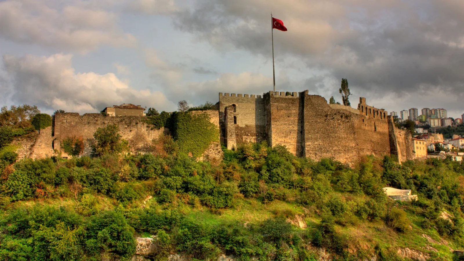 Yeşil tepelerde ortaçağ taş kalesi, Türk bayrağı, bulutlu gökyüzü.
