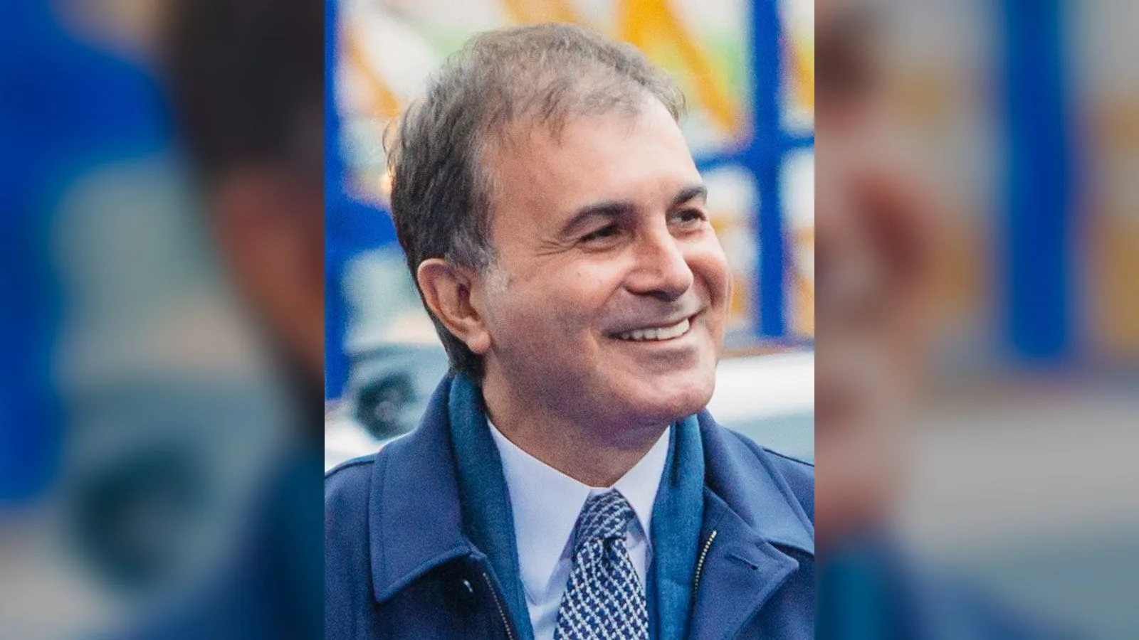 Takım elbiseli, gözlüklü orta yaşlı erkek, bulanık mavi-sarı logo arkası.