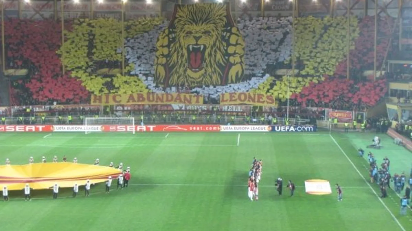 Galatasaray stadyumunda hazırlık maçında oyuncular saha ortasında duruyor, tribünde aslan ve sarı-kırmızı renklerdeki kurulu görülüyor.