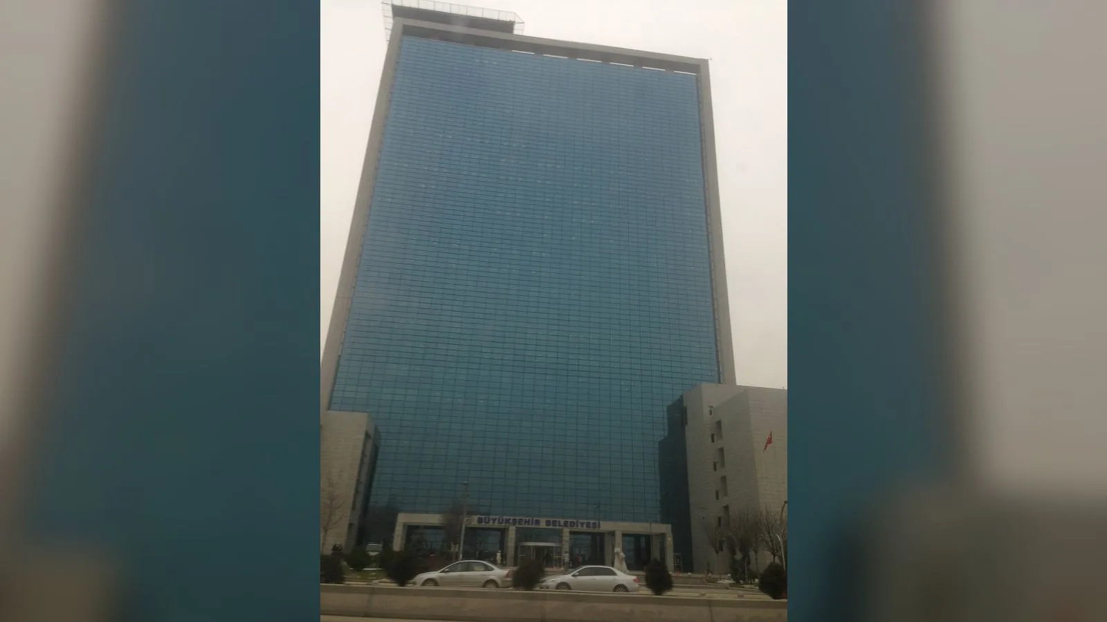 Ankara Büyükşehir Belediyesi binasının ön cephesi, iki beyaz araç ve yan bina.