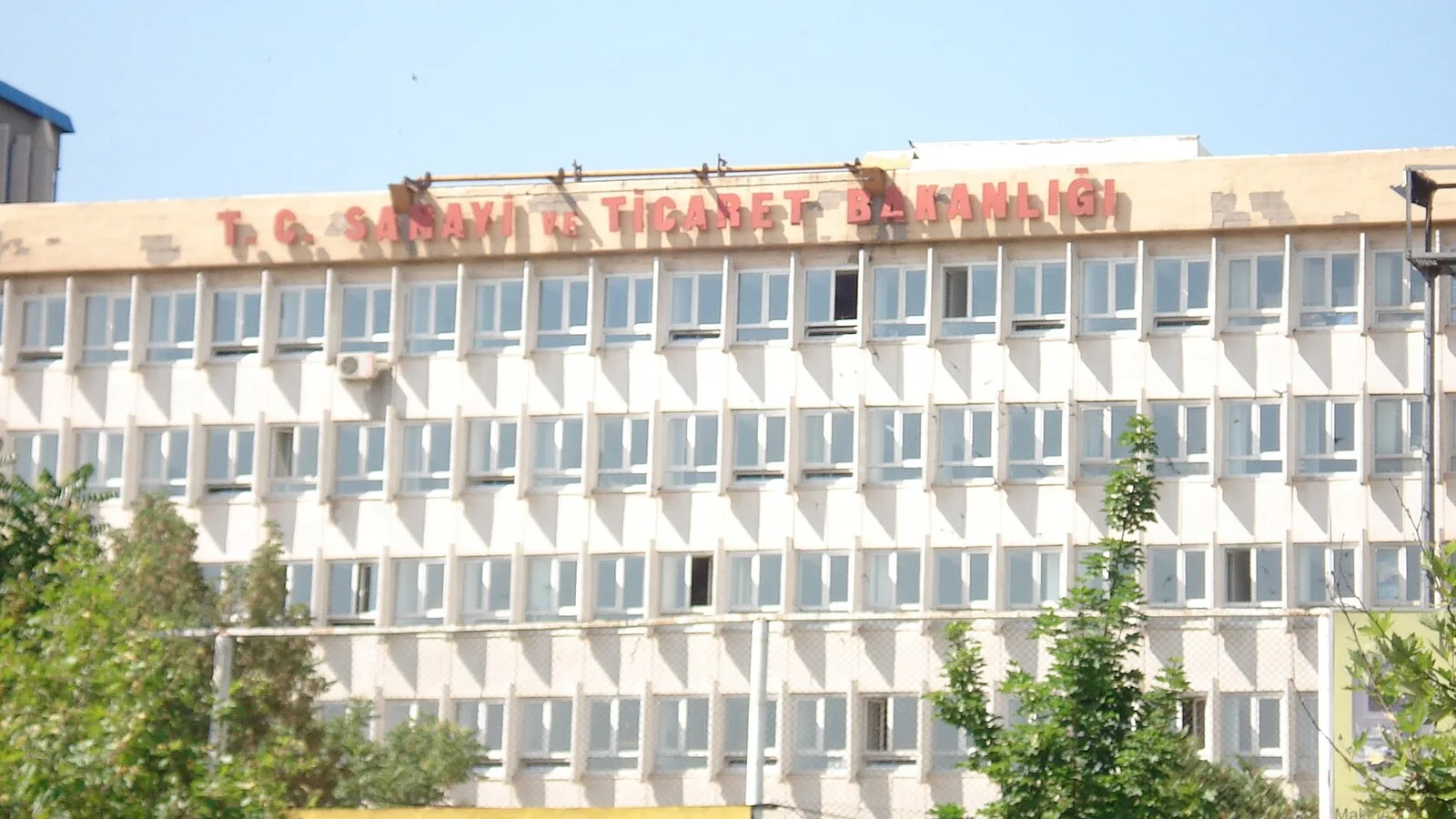 Beyaz, ağır yapılı bir ofis binasının üst kısmında kırmızı-sarı harflerle Türkçe yazı.