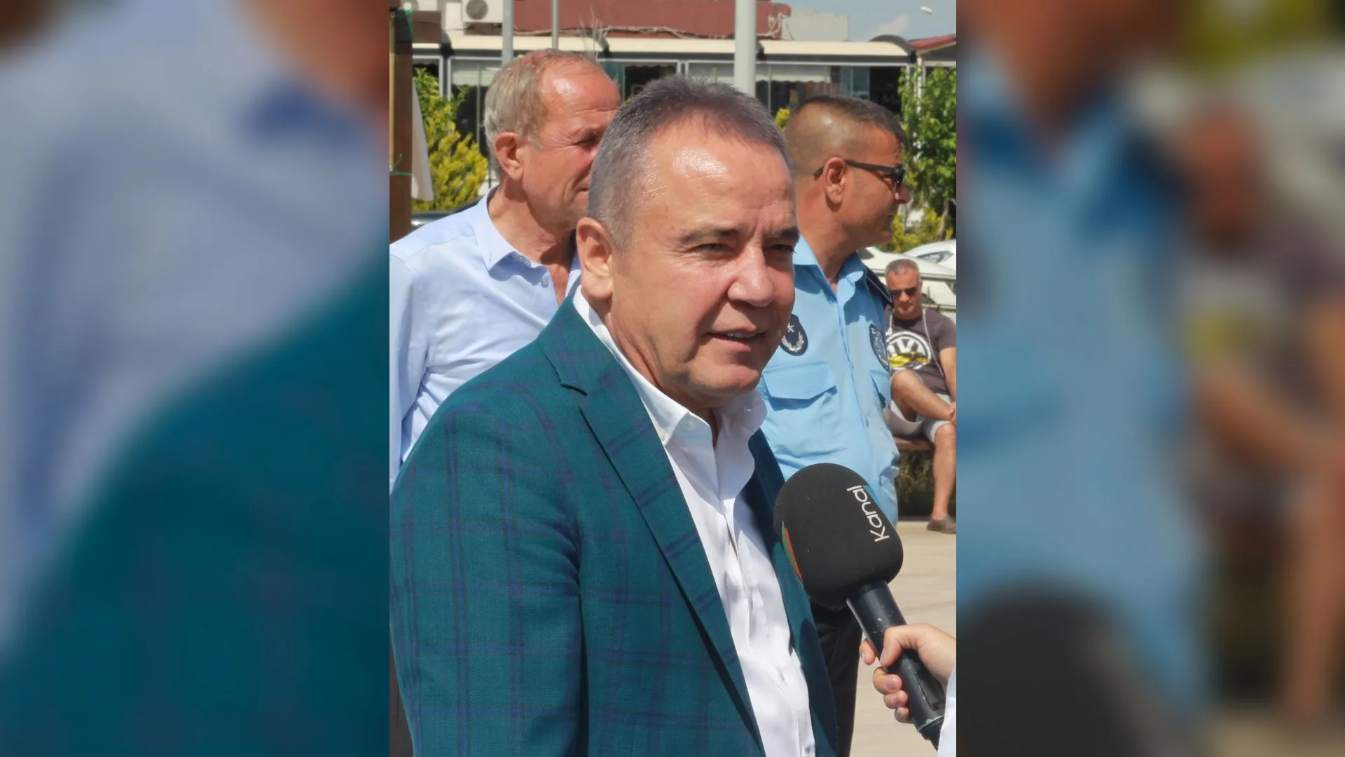 Ekose ceketli, beyaz gömlekli bir adam mikrofona konuşuyor; arkasında iki adam duruyor.