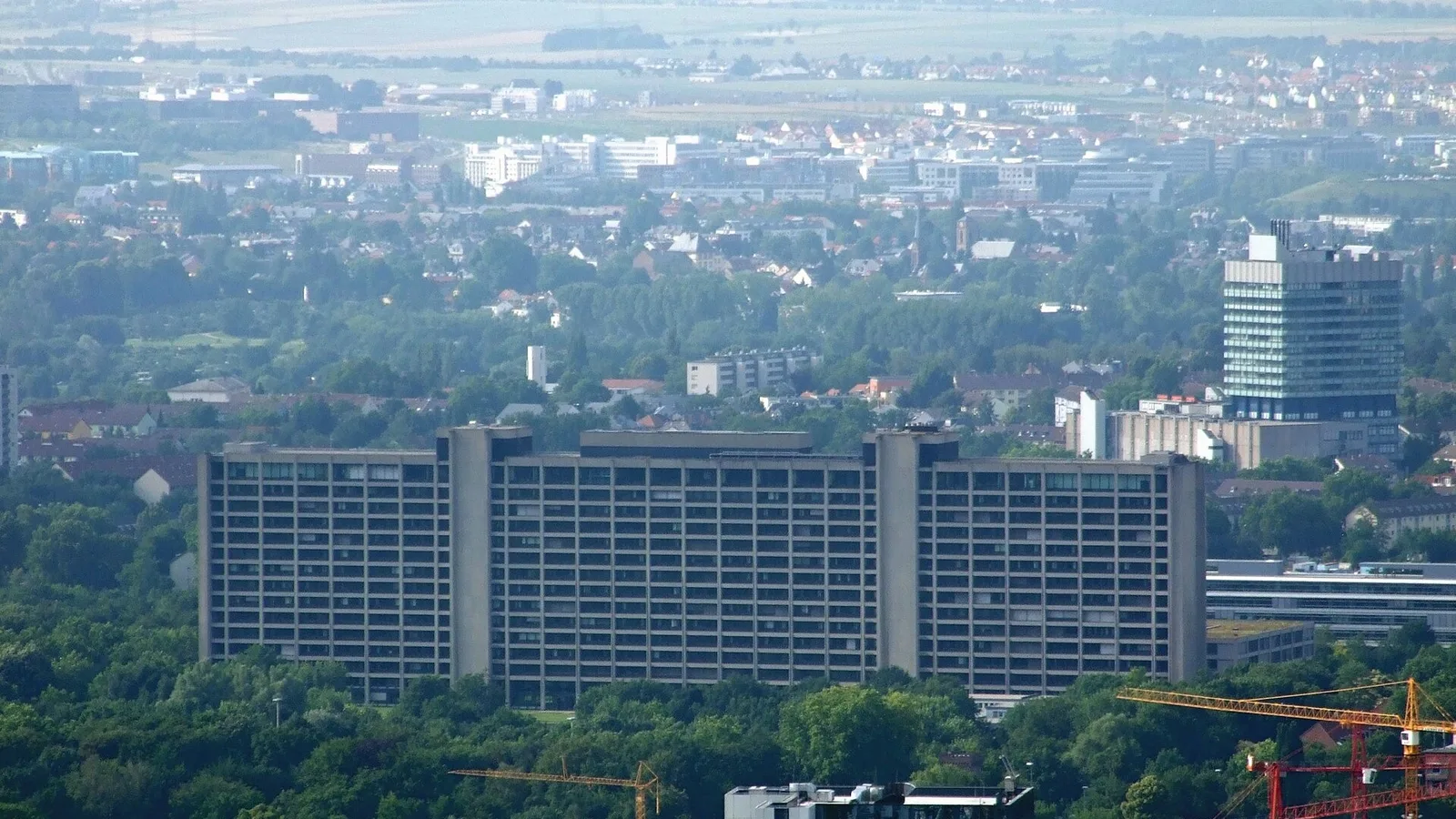 Alman şehrinin panoramik hava görüntüsü, gri bina kompleksleri ve ağaçlarla çevrili.