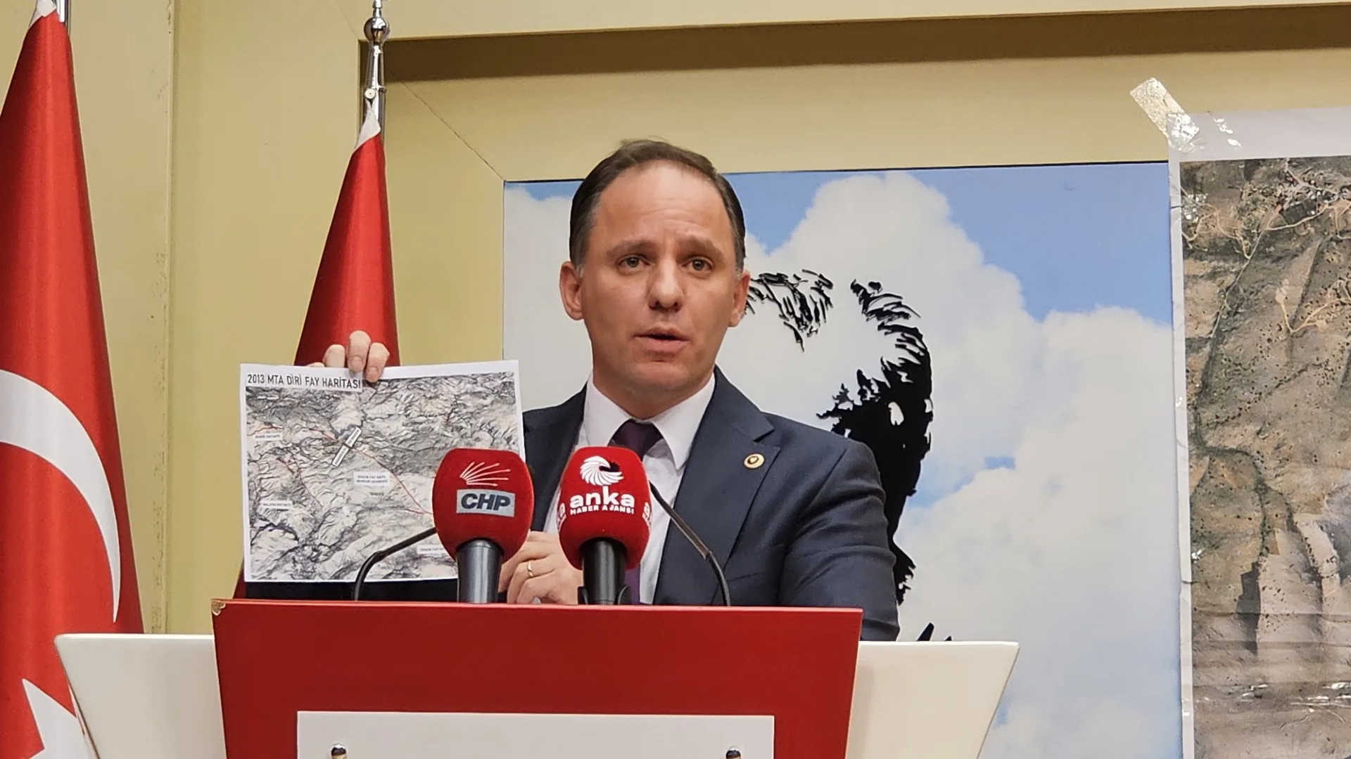 Takım elbiseli bir adam kürsüde mikrofonlar önünde elinde harita tutuyor