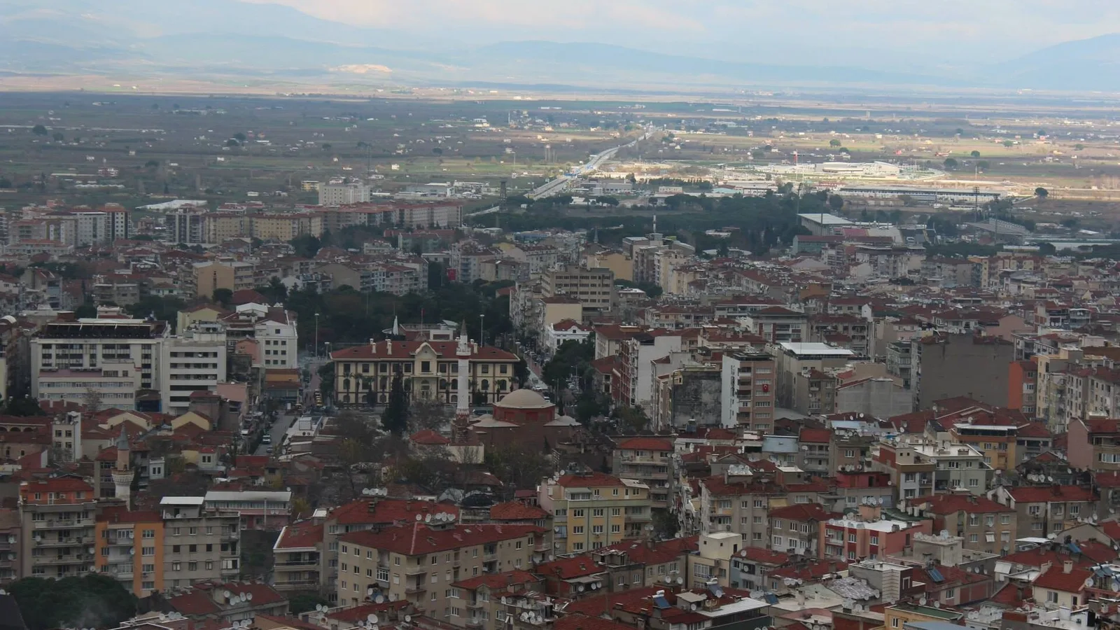 Kırmızı çatılı evler, endüstriyel alanlar ve tarım sahaları gösteren geniş şehir manzarası.