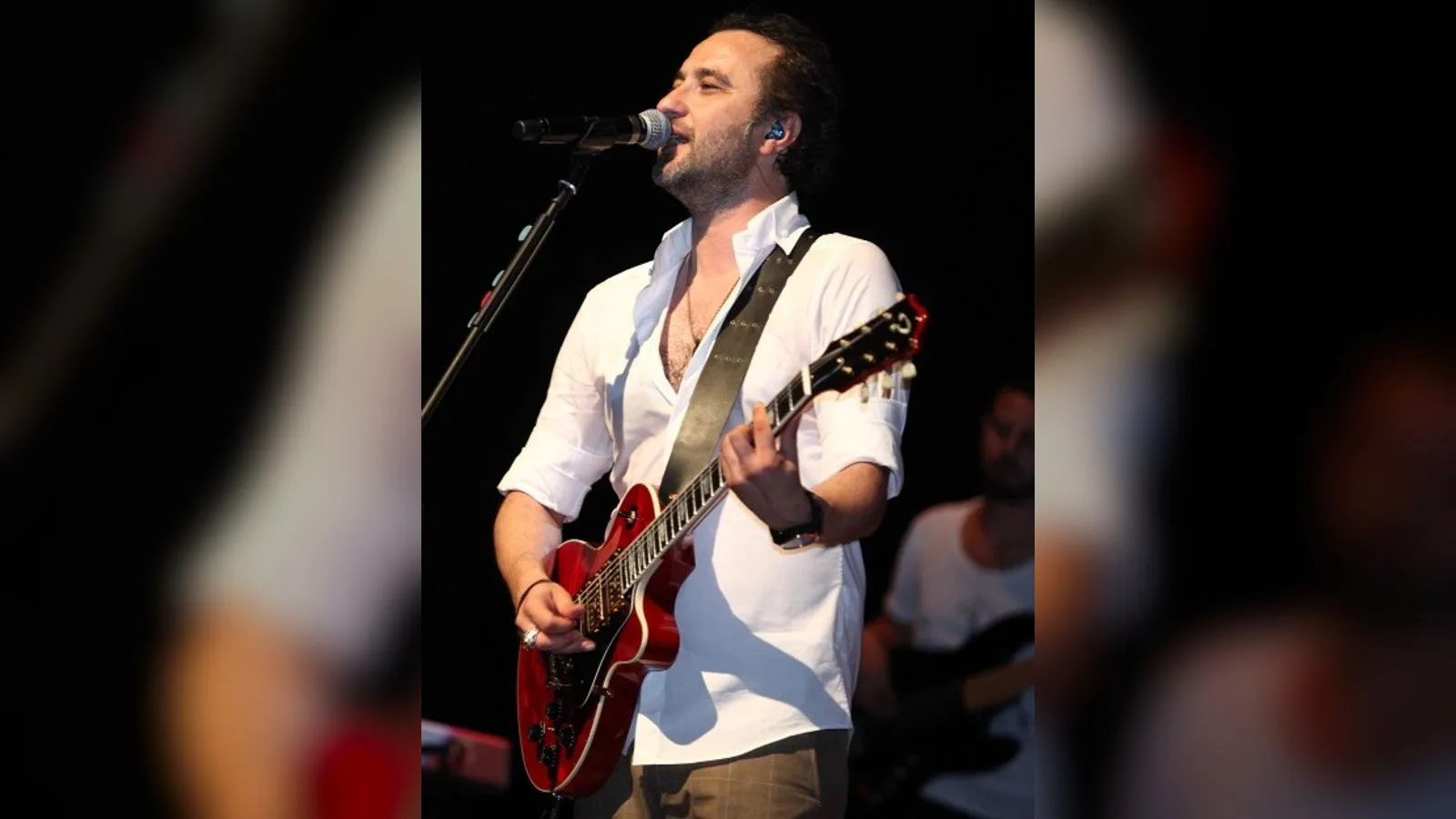 Beyaz gömlek ve kırmızı gitar takan şarkıcı, mikrofona şarkı söylüyor.