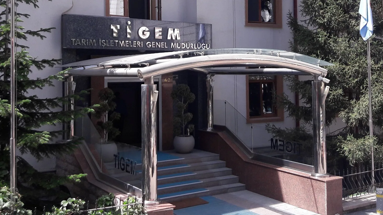 TİGEM yazısı taşıyan kurumsal binaya ait cam giriş kapısı ve merdivenleri.
