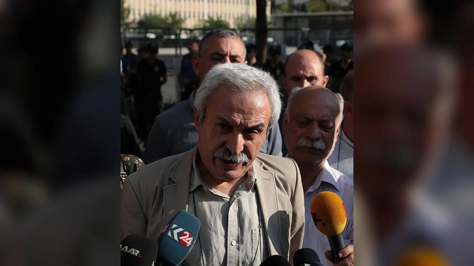Bej ceketi ve gözlüğü olan beyaz saçlı bir erkek mikrofon önünde konuşurken.