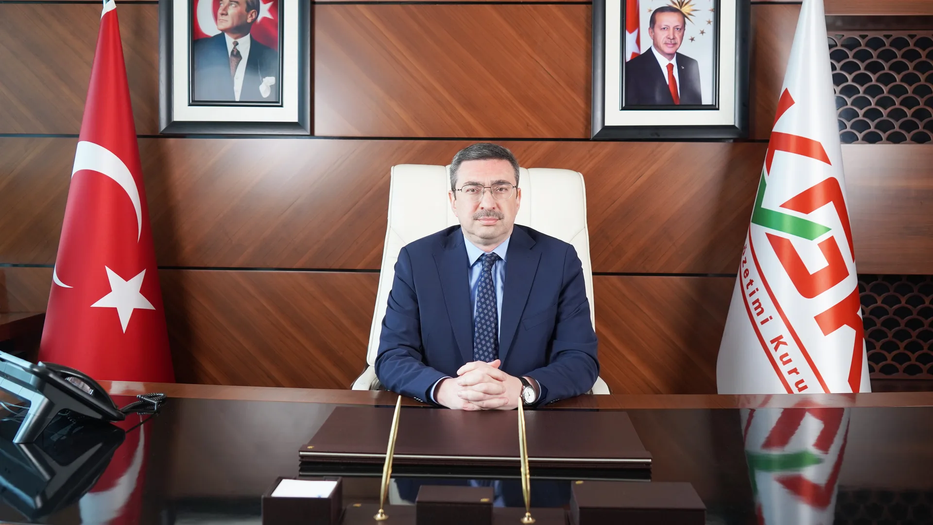 Bayraklar ve portreler önünde makam masasında oturan takım elbiseli gözlüklü adam