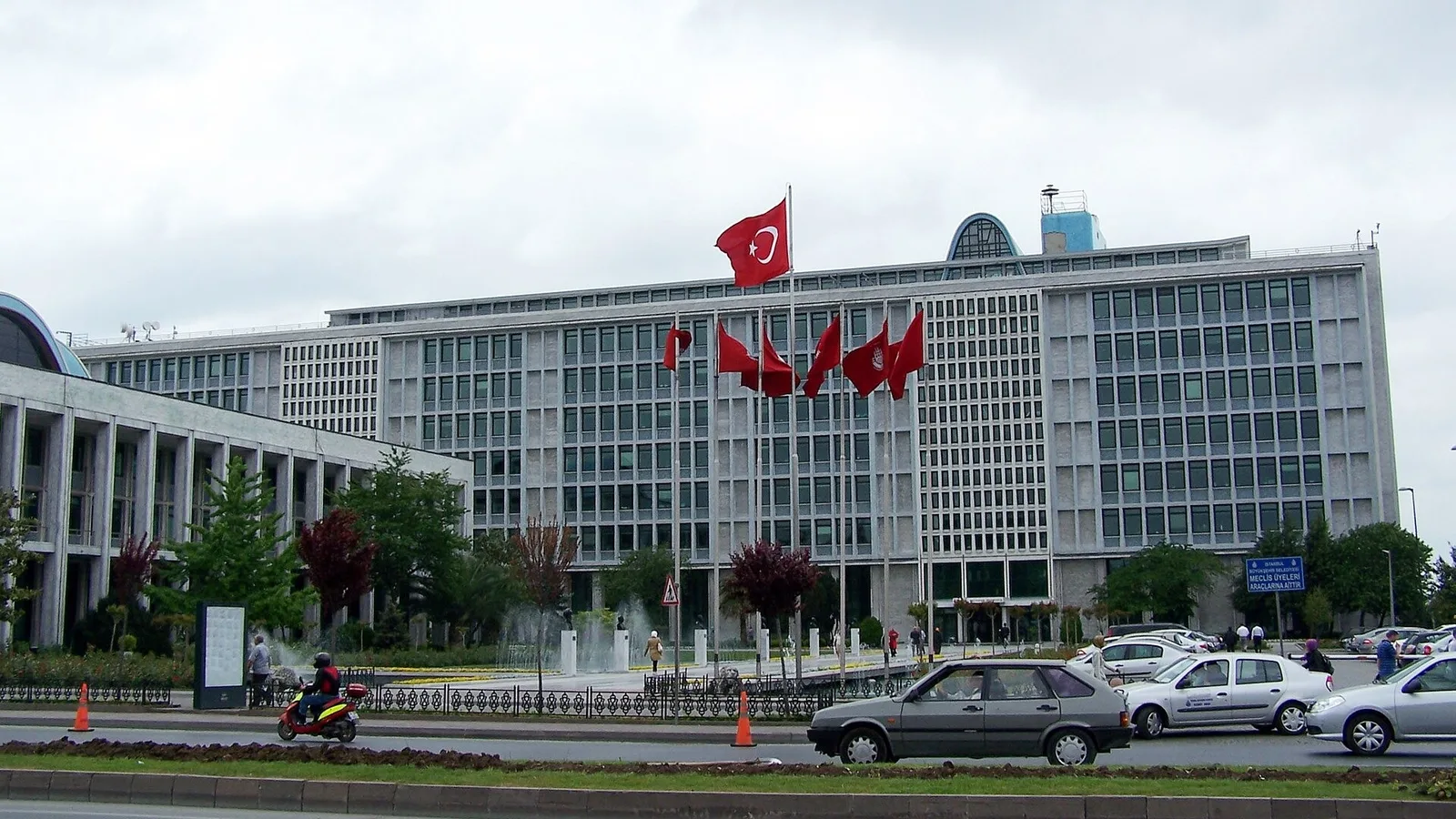 Bayraklı gri cepheli beş katlı resmi bir bina, ön tarafında polis hizmet araçları ve bariyerler.