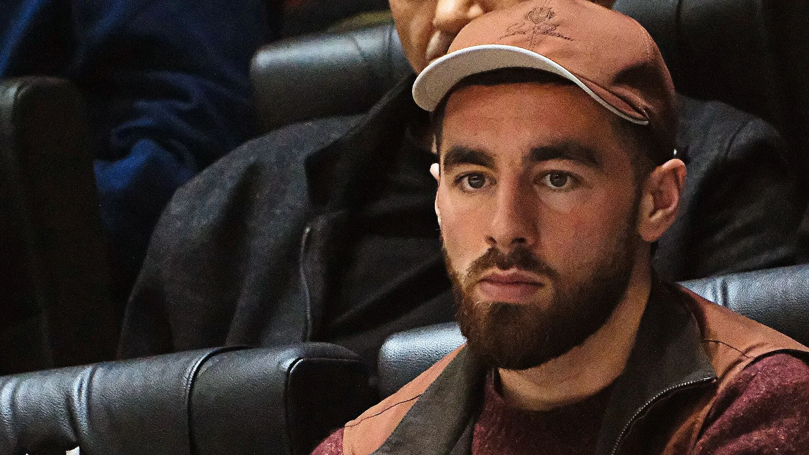 Bearded man in brown cap sitting on bench, wearing burgundy jacket.