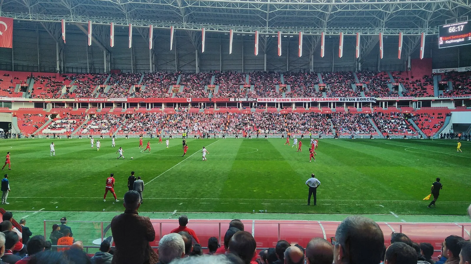 Kırmızı ve beyaz formalı oyuncuların oynadığı, kalabalık tribünlü bir futbol stadyumu.