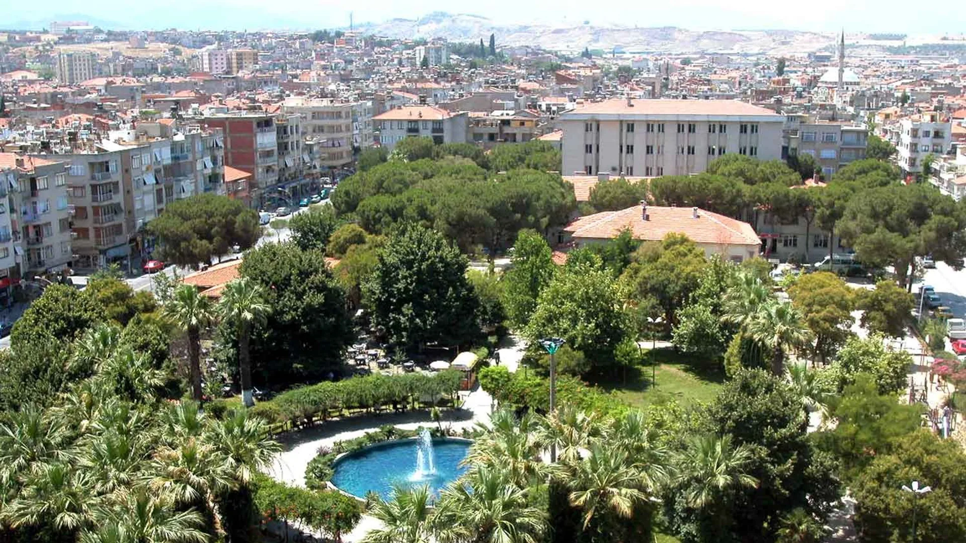 Turgutlu şehrinin panoramik manzarası, ön planda yeşil alan ve yüzme havuzu, arka planda apartmanlar ve dağlar.