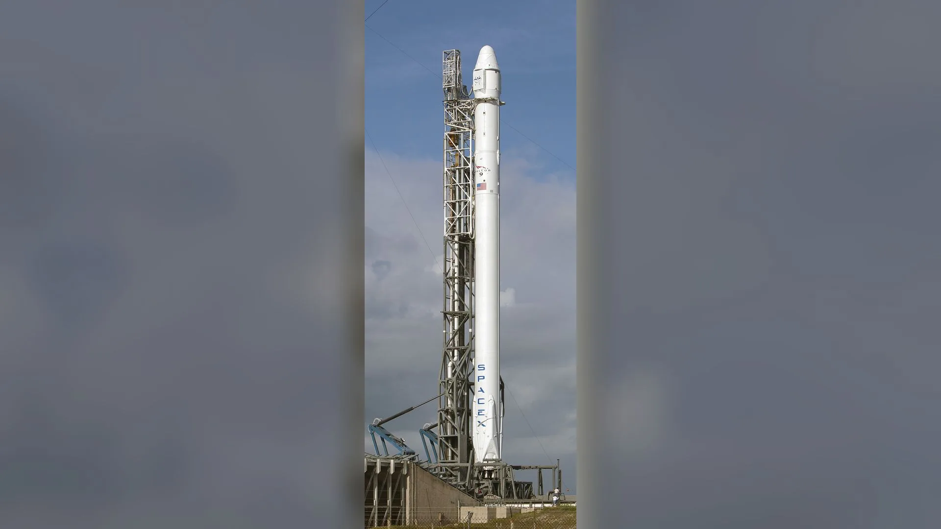 Rampa kulesinin yanında dikey duran, üzerinde SpaceX yazan beyaz roket, arka planda bulutlu gökyüzü