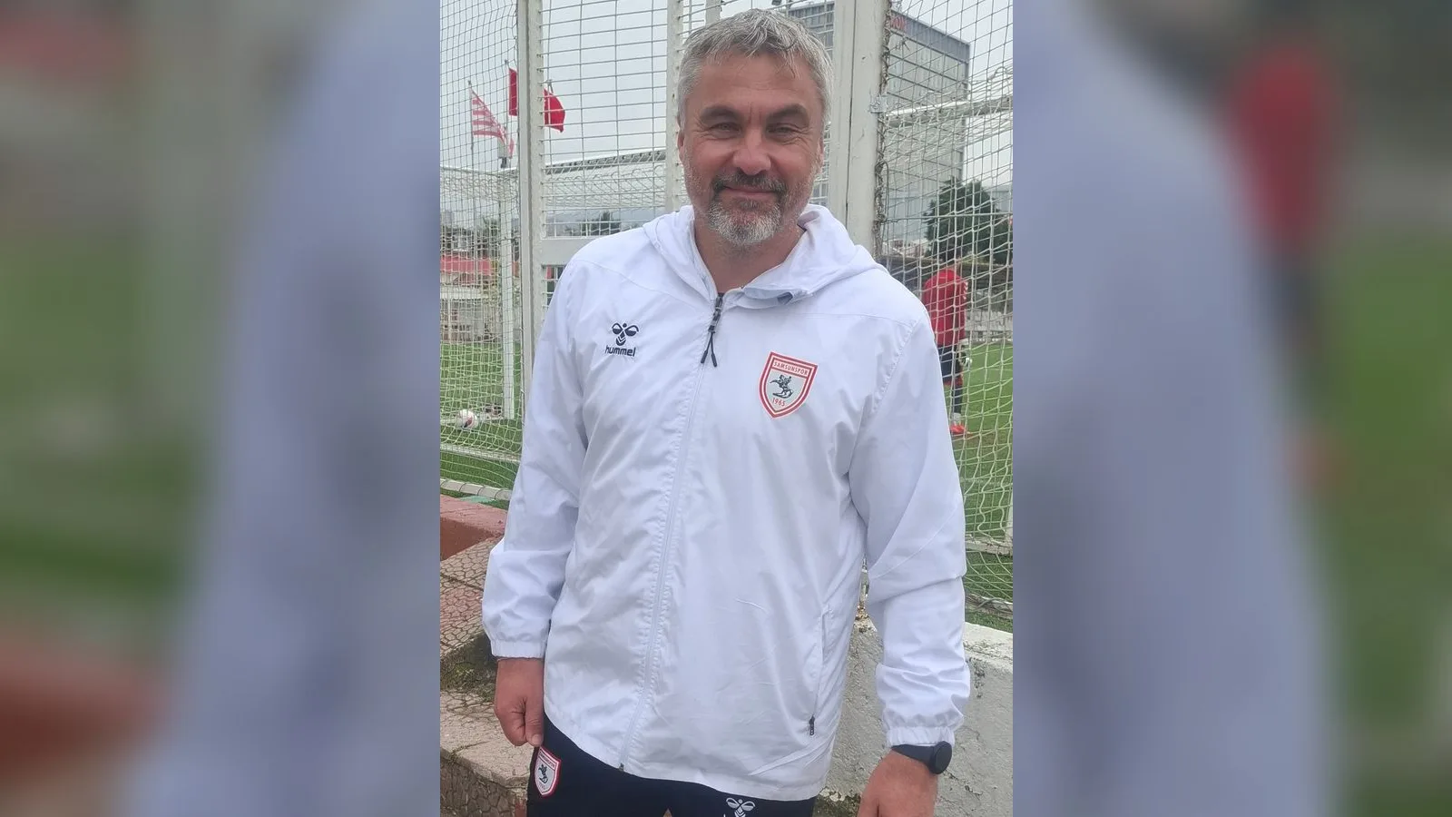 Beyaz antrenman ceketi giymiş, gri saçlı erkek, futbol sahasında poz veriyor.