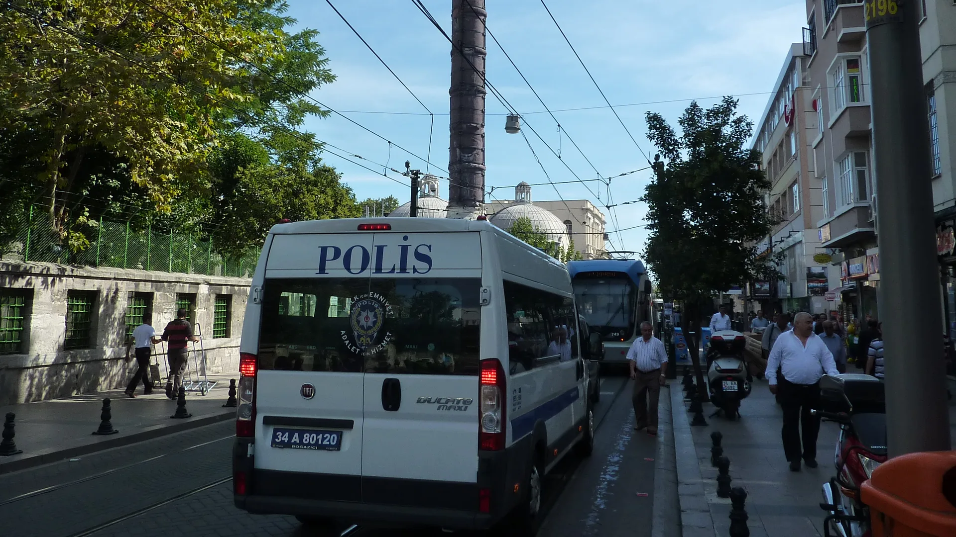 Tarihi taş sütun önünde duran beyaz polis minibüsü ve tramvay, kaldırımda yürüyen insanlar