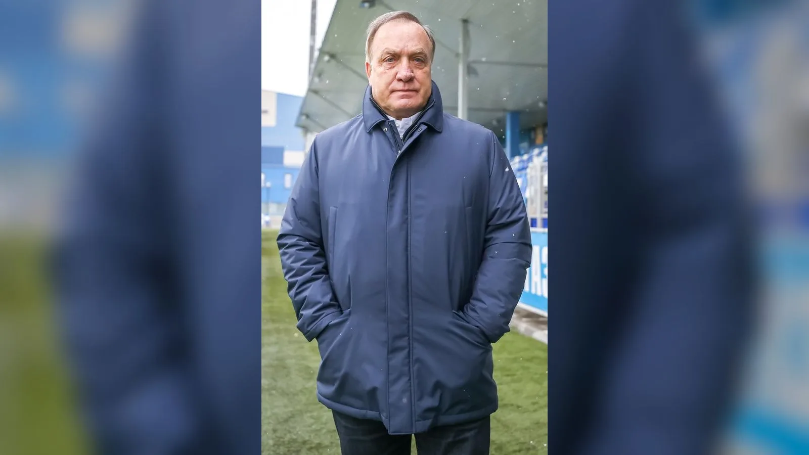 Orta yaşlı, lacivert ceketi giymiş bir adam futbol stadyumunun önünde durmuş.