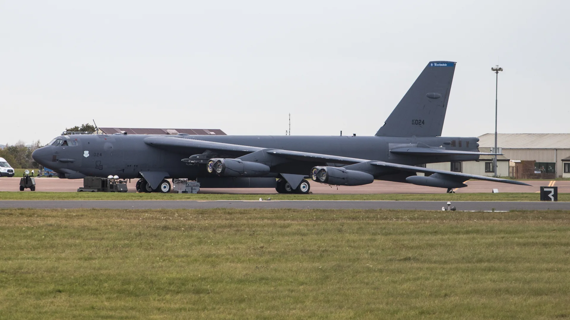 Pistin yanında park etmiş gri renkli büyük bir B-52 bombardıman uçağı ve arkada hangarlar.