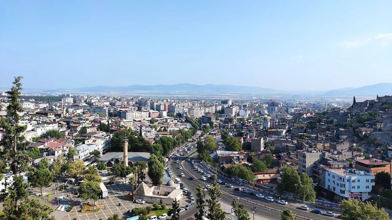 Tepesi yüksek bir tepenin üzerinden şehir manzarası: çok katlı binalar, ağaçlar, geniş ana cadde ve dağlık ufuk görülüyor.