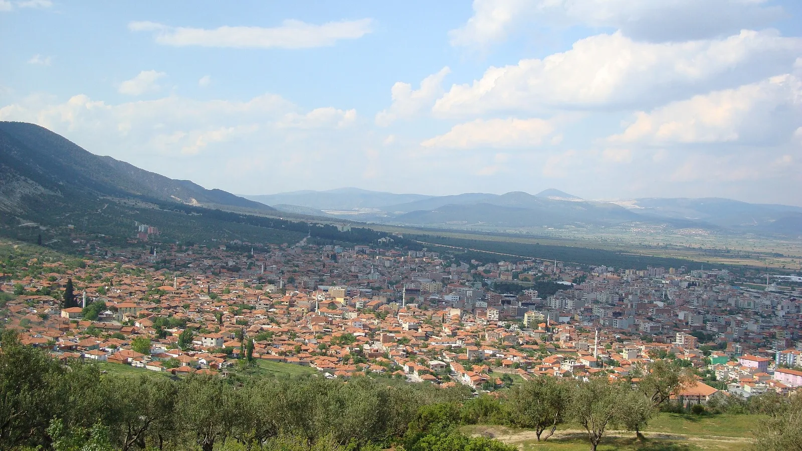 Yeşil dağlarla çevrili, turuncu çatılı evlerin sıralandığı şehir manzarası.