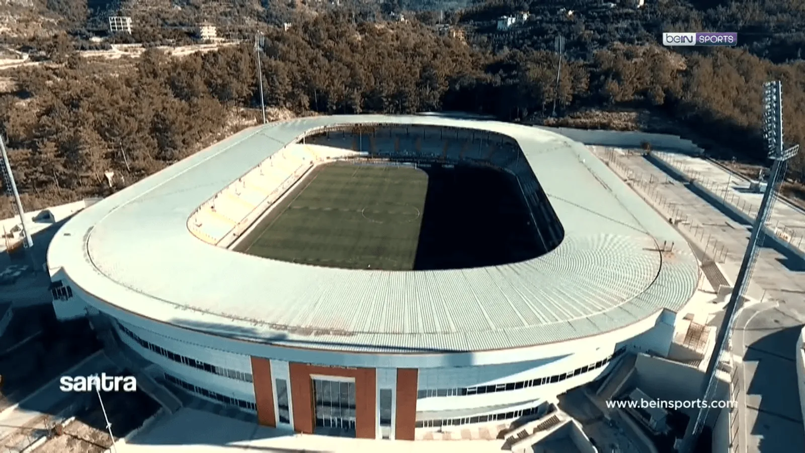 Alanya şehri ve ormanlı arkaplanın üzerinde beyaz çatılı futbol stadyumu.