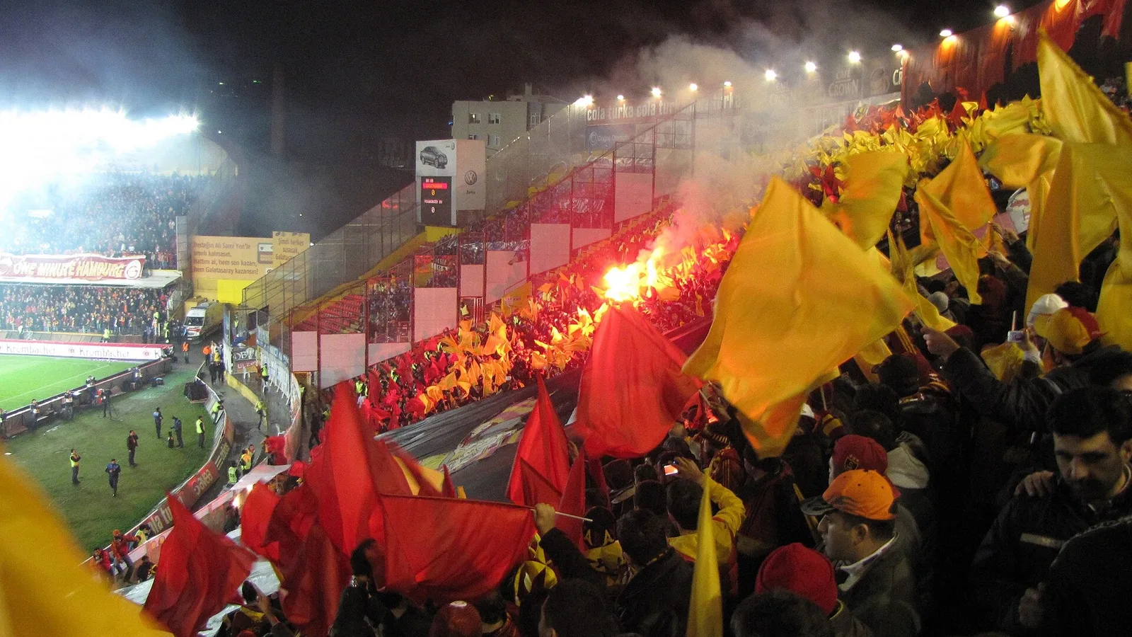 Stadyumda kırmızı-sarı bayraklar ve alevli tezahürat, futbol maçı.