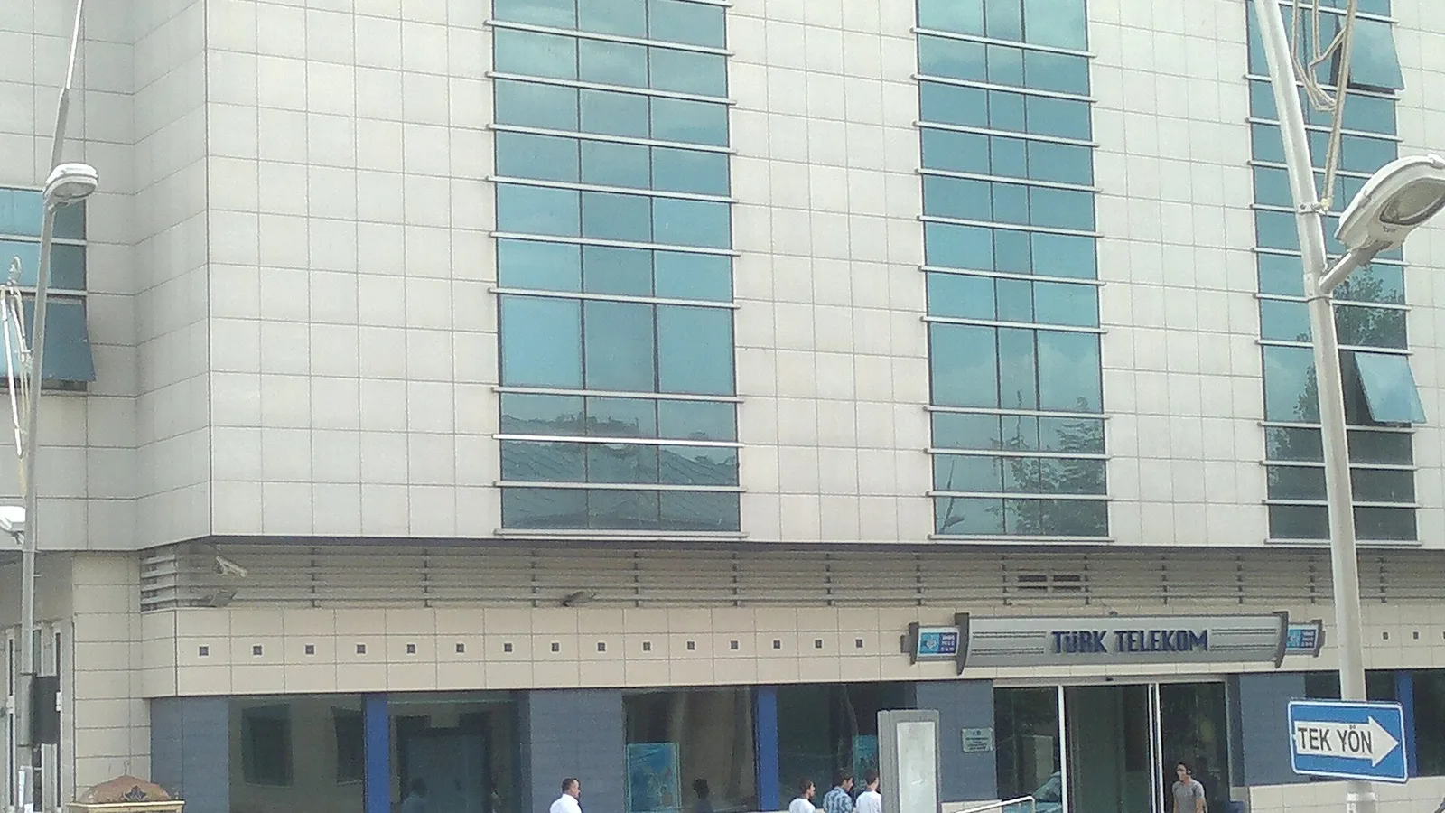 Türk Telekom mağazasının ön cephesi, beyaz ve cam panellerle kaplı, sokakta yürüyen insanlar.