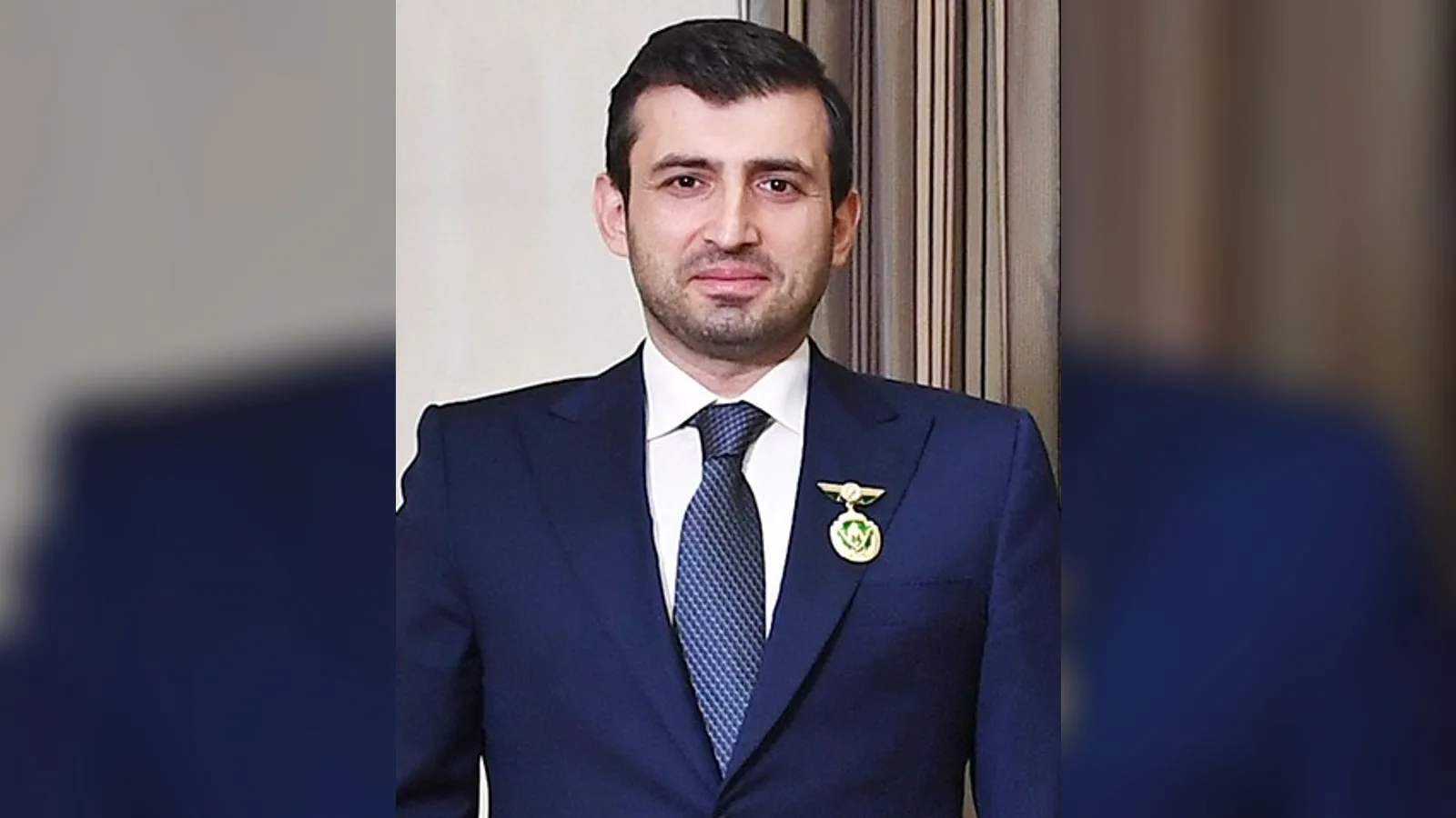 Takım elbiseli, mavi ceketli, boynunda lacivert kravatı ve göğsünde altın rozetli bir adam.
