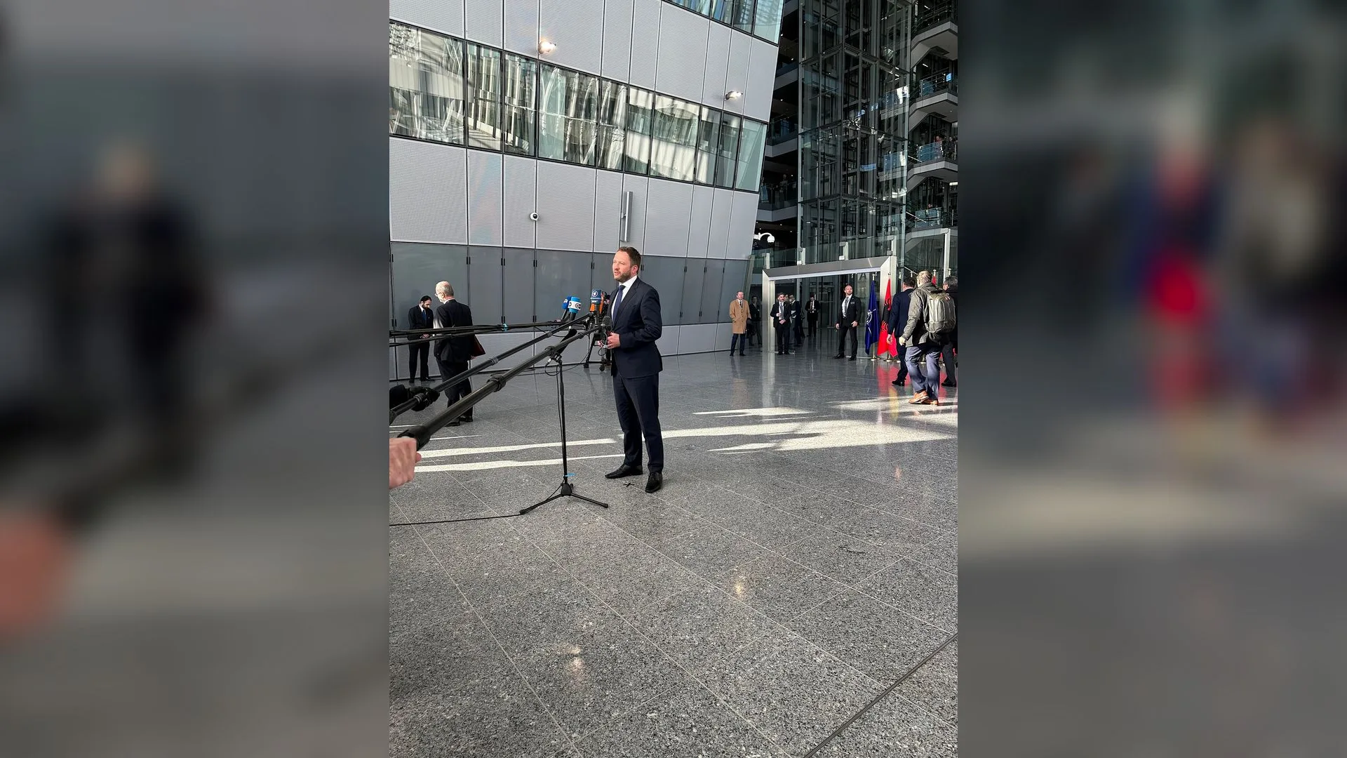 Takım elbiseli bir adam cam cepheli binanın holünde mikrofonlara konuşuyor