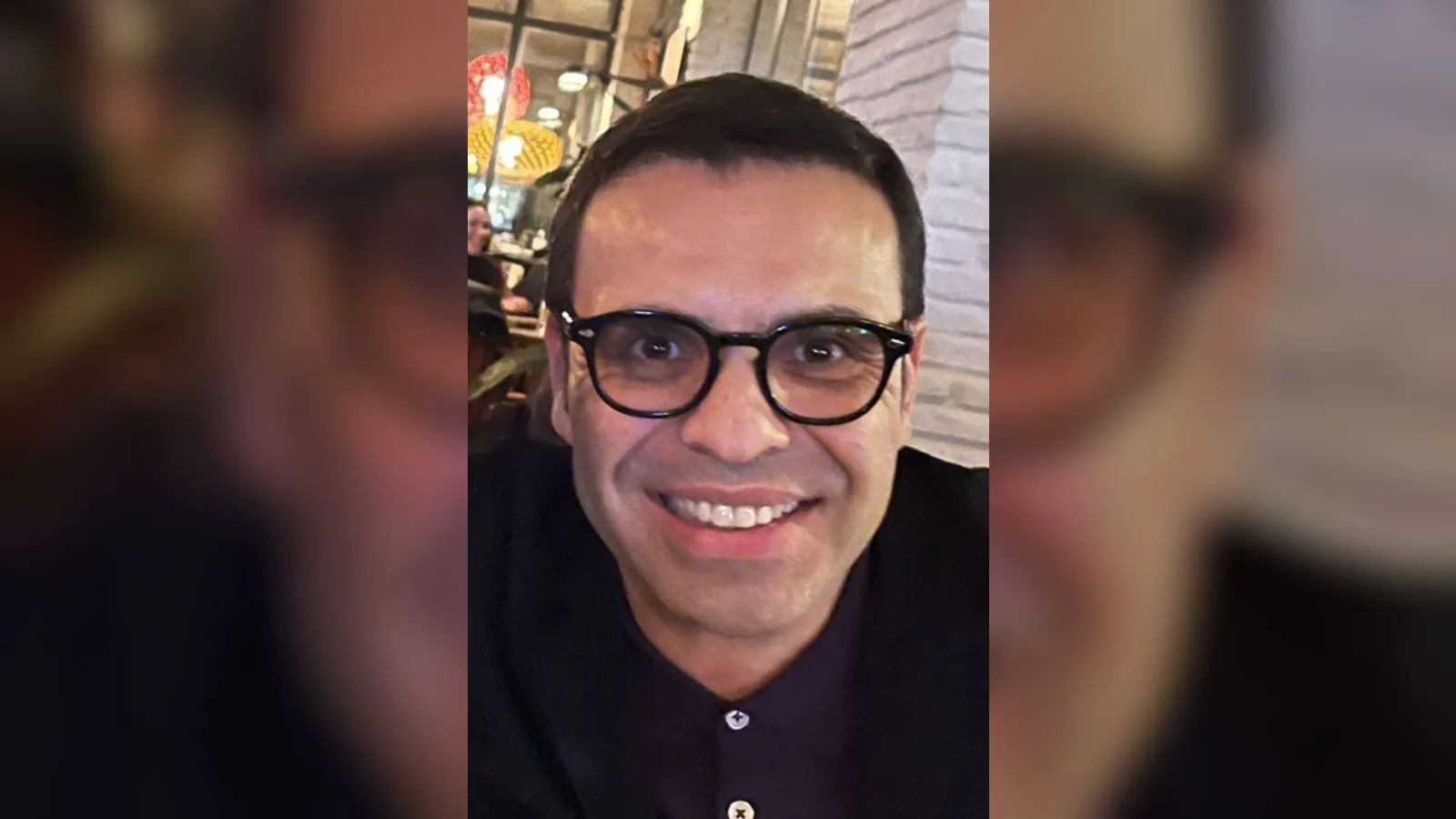 Gözlüklü, siyah gömlek giymiş bir adam, resepsiyon alanında gülümsüyor.
