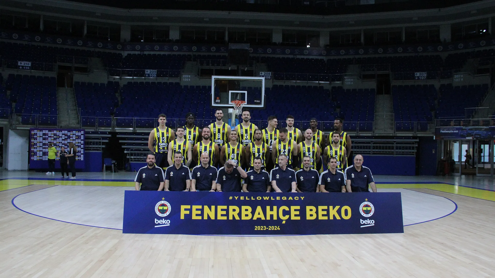Sarı-lacivert formalı basketbolcular ve teknik ekip, önlerinde Fenerbahçe Beko pankartıyla salonda poz veriyor.