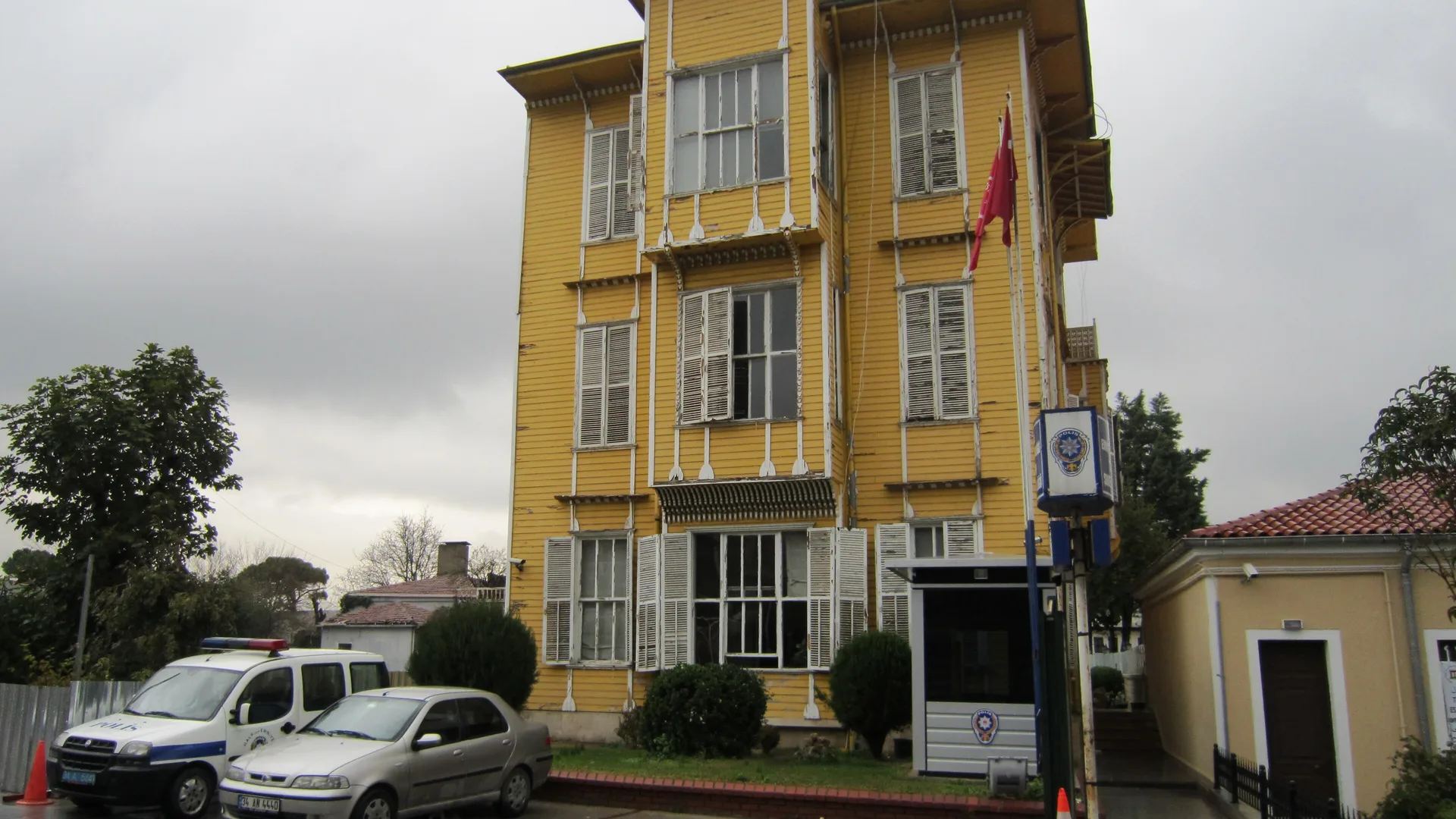 Bulutlu havada Türk bayrağı asılı sarı ahşap bir karakol binası, önünde polis aracı
