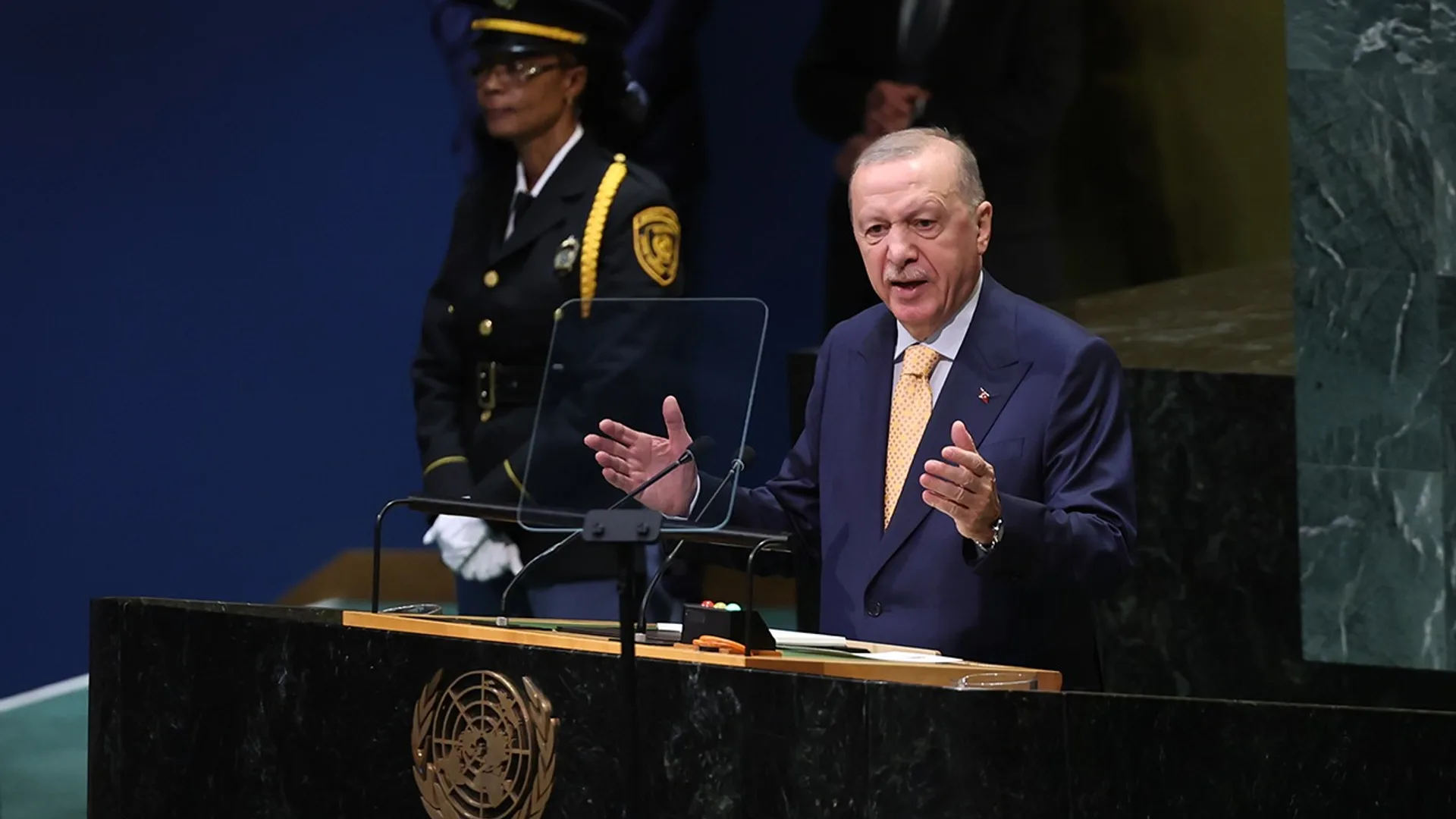 Cumhurbaşkanı Recep Tayyip Erdoğan, Birleşmiş Milletler Genel Kurulu kürsüsünde el hareketleriyle konuşurken; kürsünün ön yüzünde BM amblemi görünüyor.