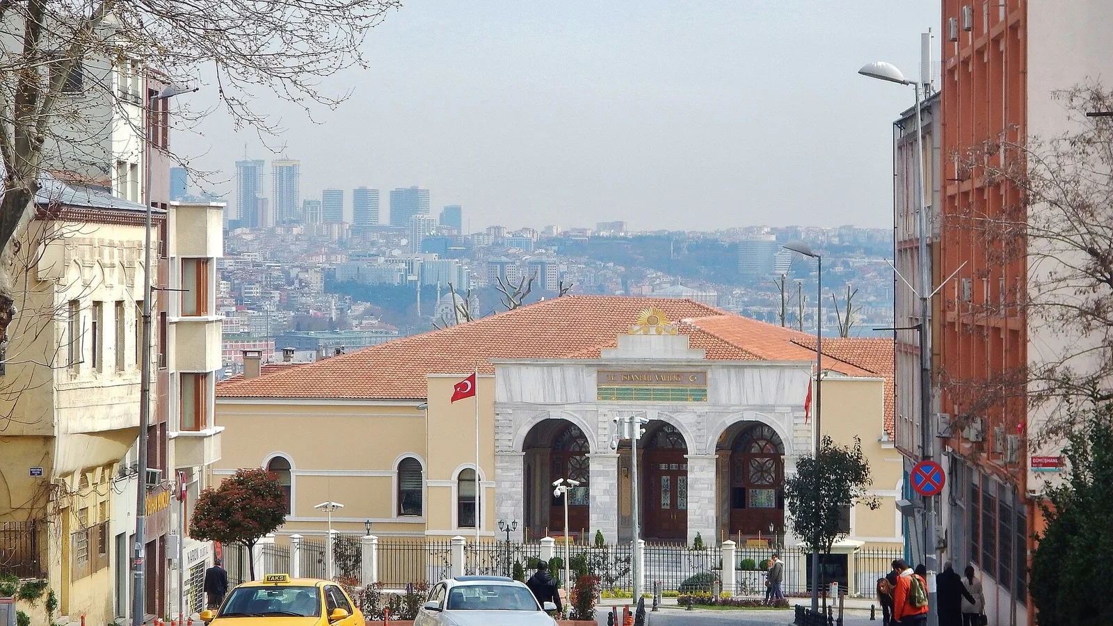 Balkon railing arkasında meydana açılan beyaz yapı, kızılcıkart çatılı, iki sarı taksi önünde.