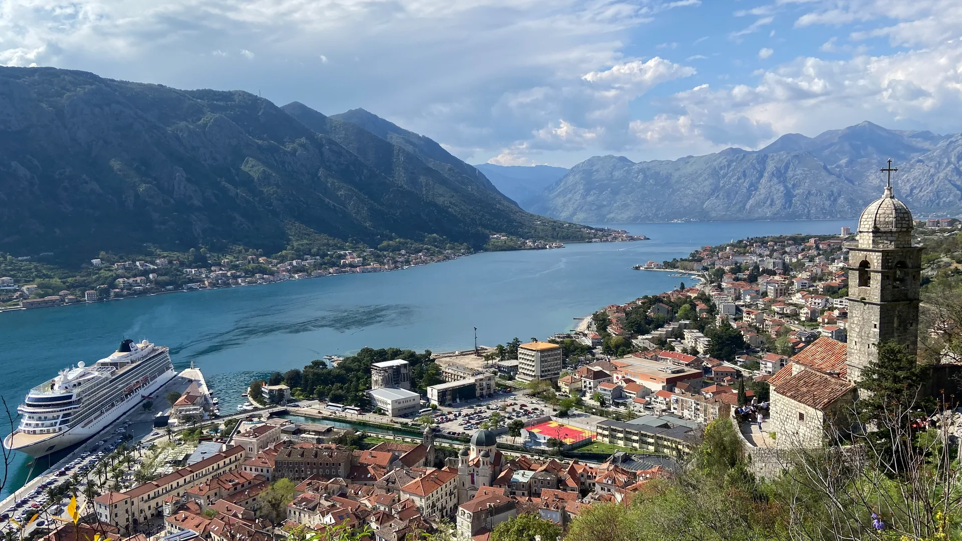 Dağlarla çevrili koyun kıyısında kırmızı çatılı eski kent, limanda yolcu gemisi ve taş çan kulesi