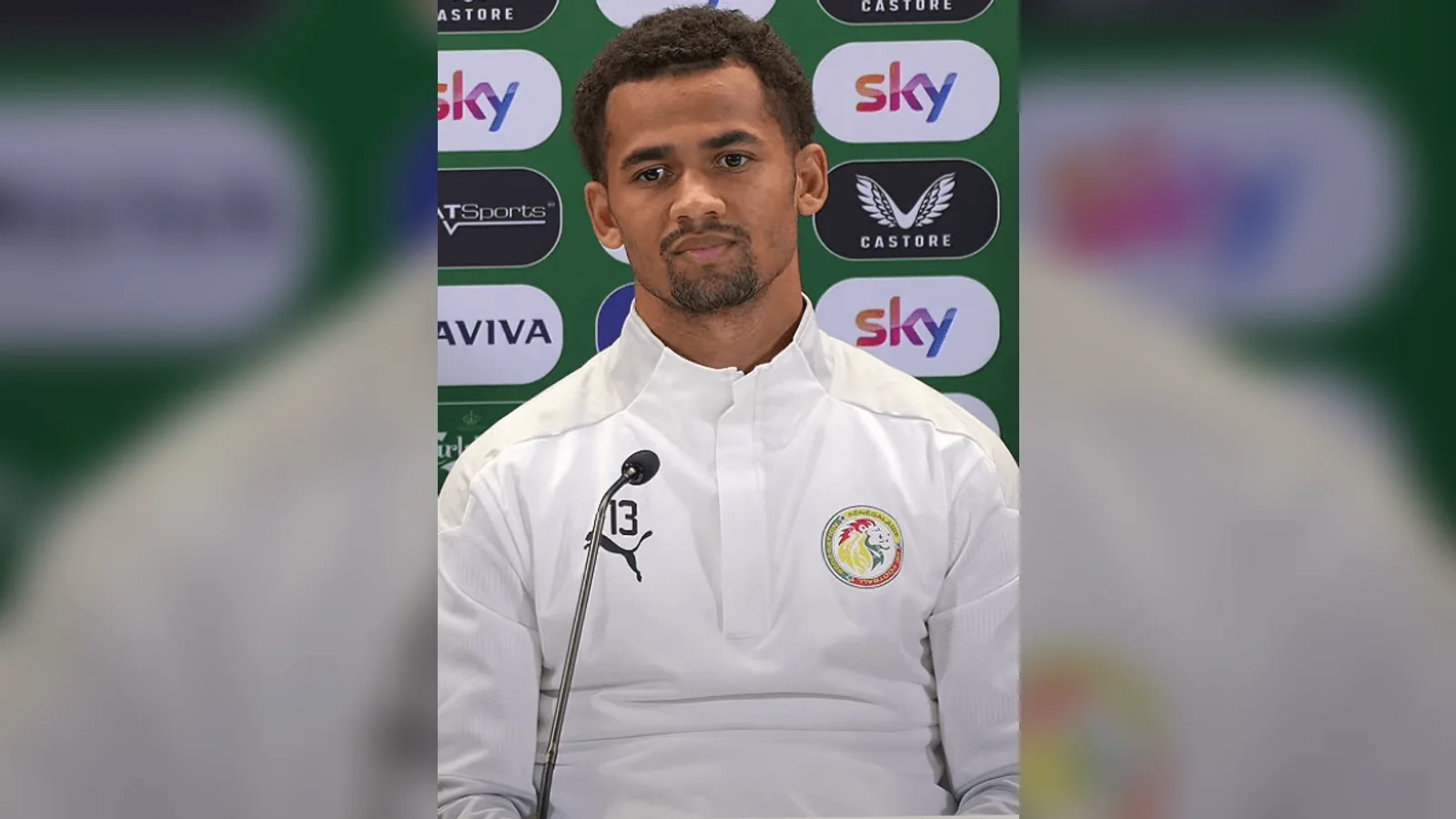 Beyaz eğitim kıyafetinde, mikrofon karşısında duran futbolcu.