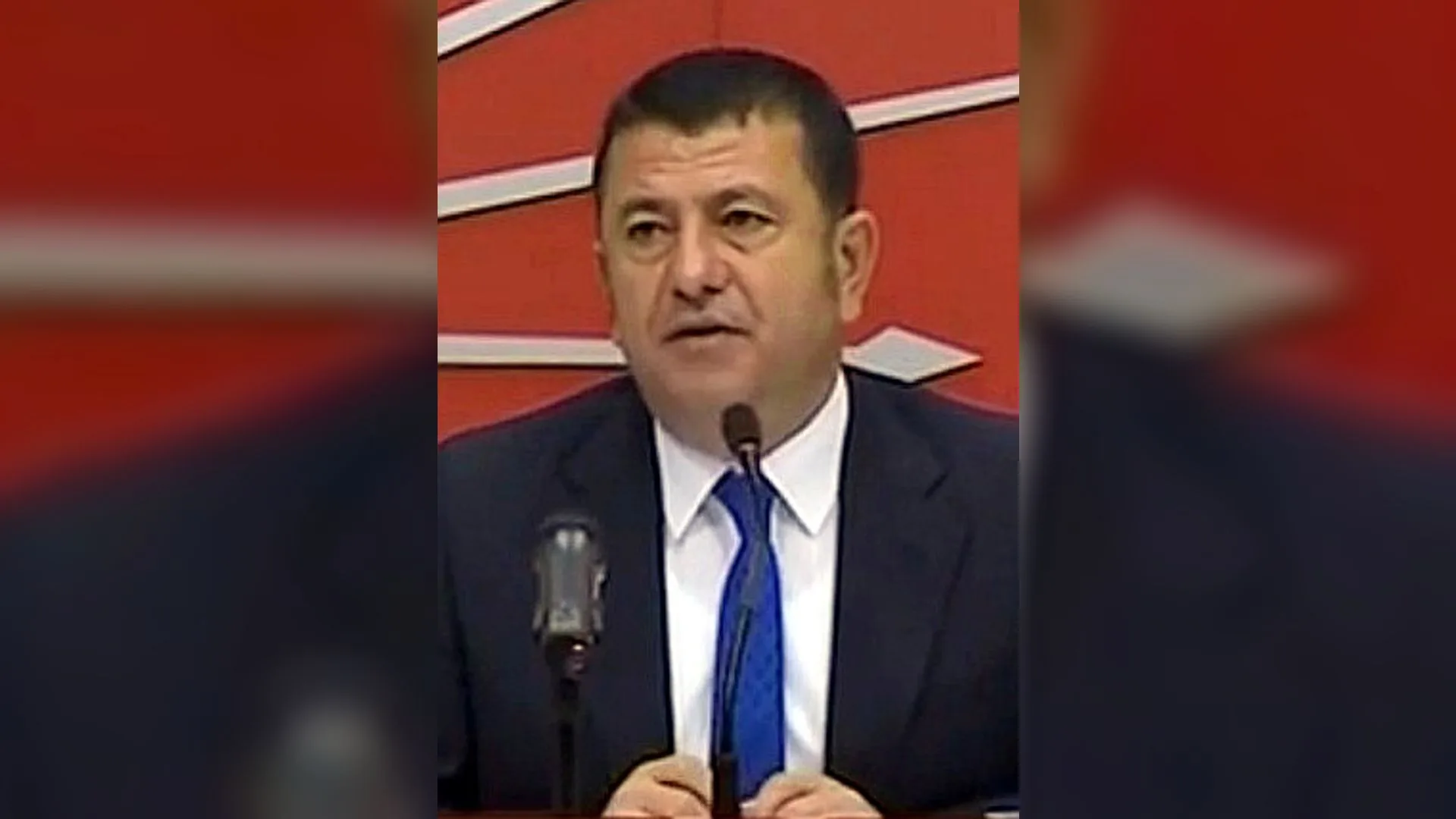 Kırmızı zemin önünde mikrofonla konuşan, lacivert takım elbiseli ve mavi kravatlı bir adam