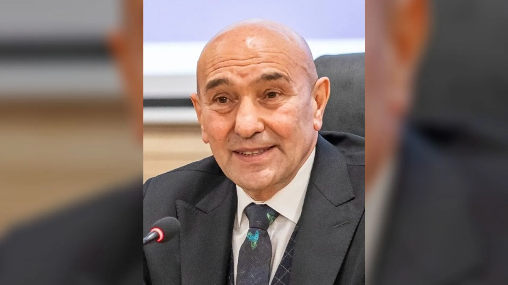 Takım elbiseli, kel, gülümseyen bir adam mikrofon önünde konuşuyor.