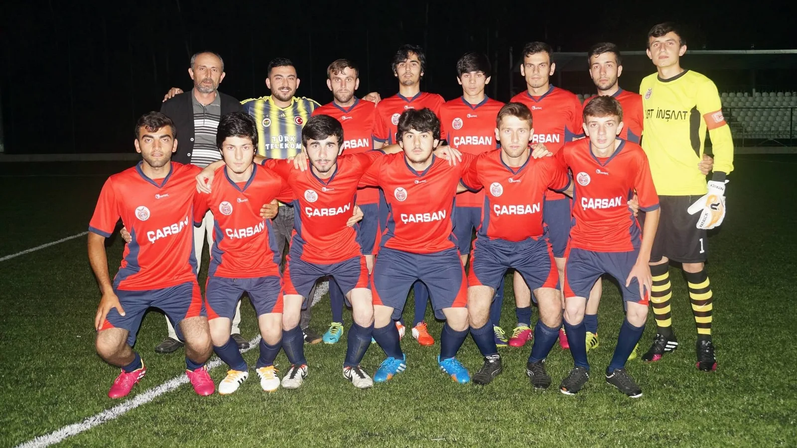 Futbol sahası, kırmızı-mavi üniforması oyuncular, sarı eldiveli kaleci ve teknik heyet.