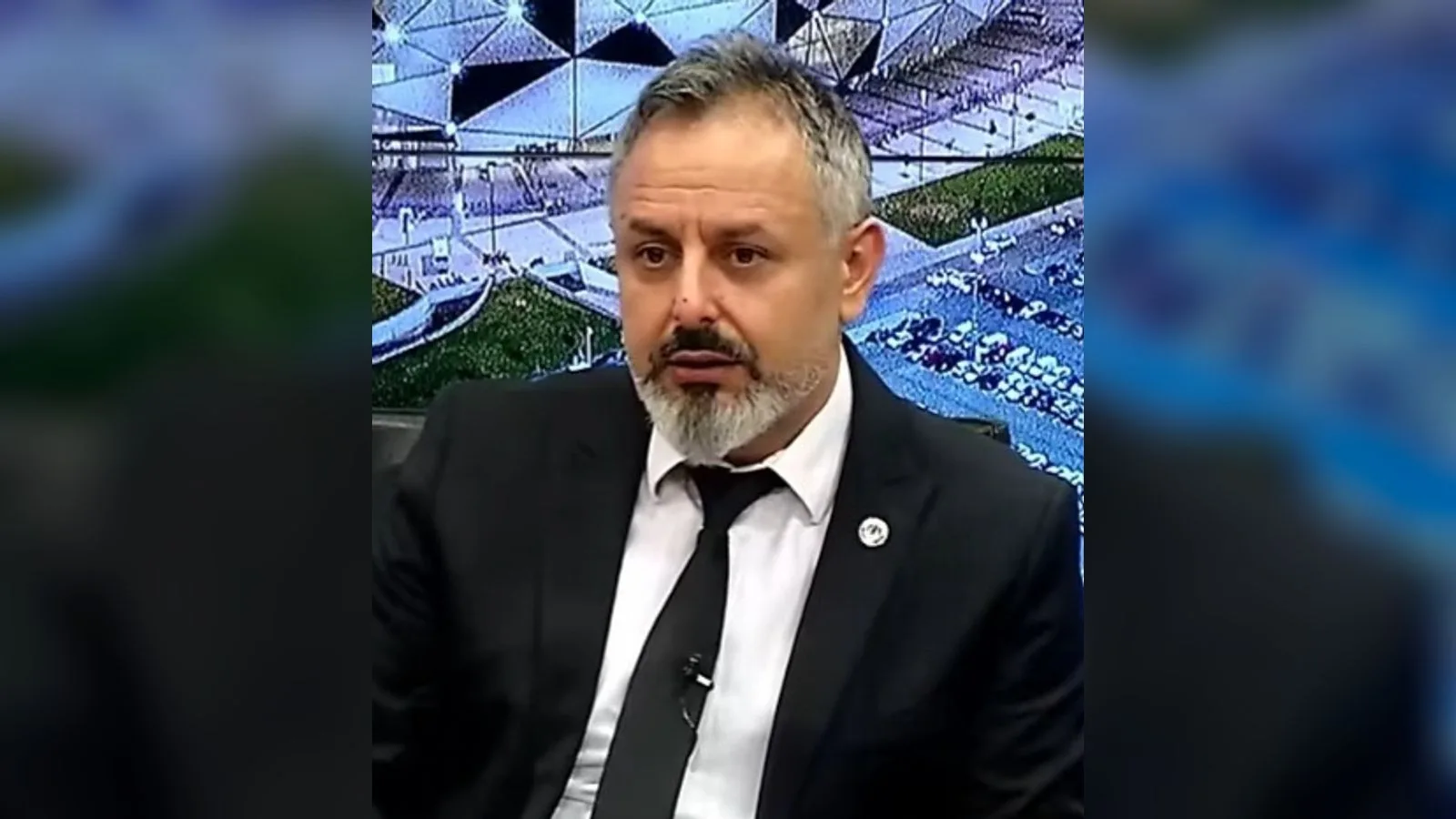 Stadyum arka planında bulunan, takım elbiseli ve kravatı giymiş, sakal olan bir adam.