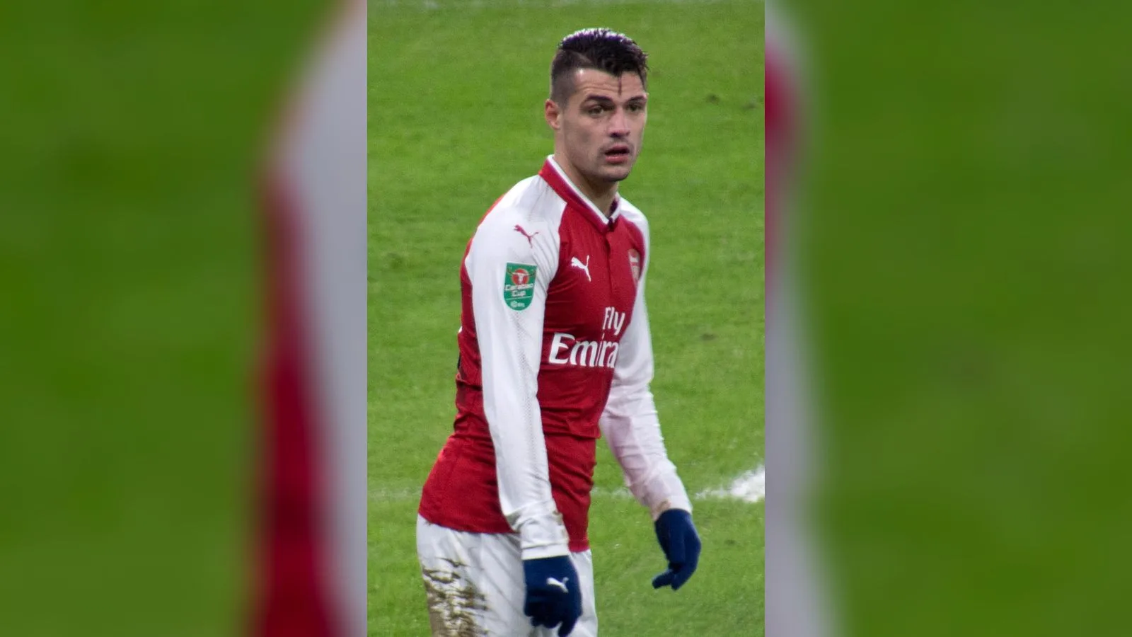 Kırmızı-beyaz formali futbolcu, çim sahada durma pozisyonunda.