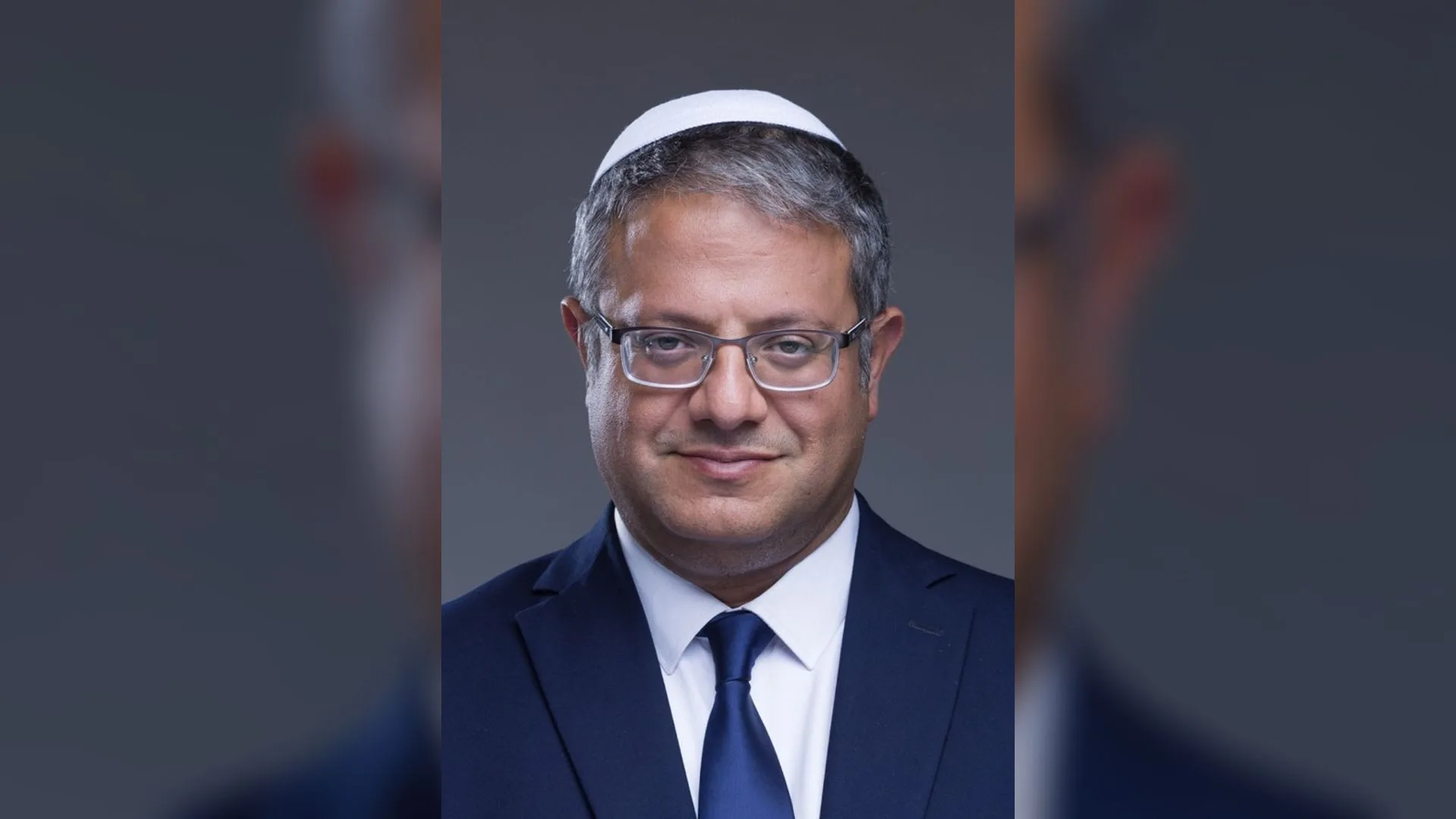 Beyaz takke ve gözlük takan, lacivert takım elbiseli ve mavi kravatlı gülümseyen bir adamın portresi