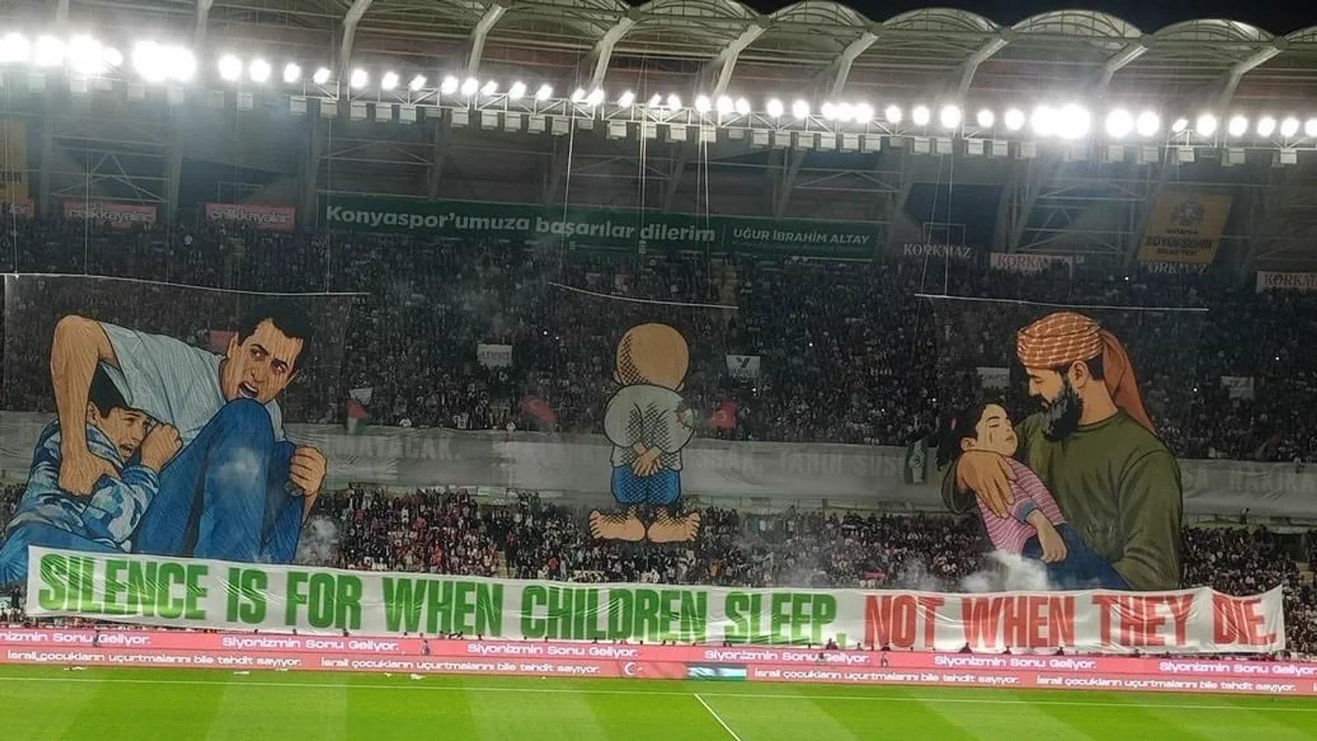 Konya Büyükşehir Stadyumu'nda tribünde açılan koreografi: solda Muhammed Durra ve babası, ortada Hanzala, sağda torununu kucağında taşıyan Halid Nebhan; altında "Silence is for when children sleep. Not when they die." yazılı pankart, sahada beyaz güvercin.