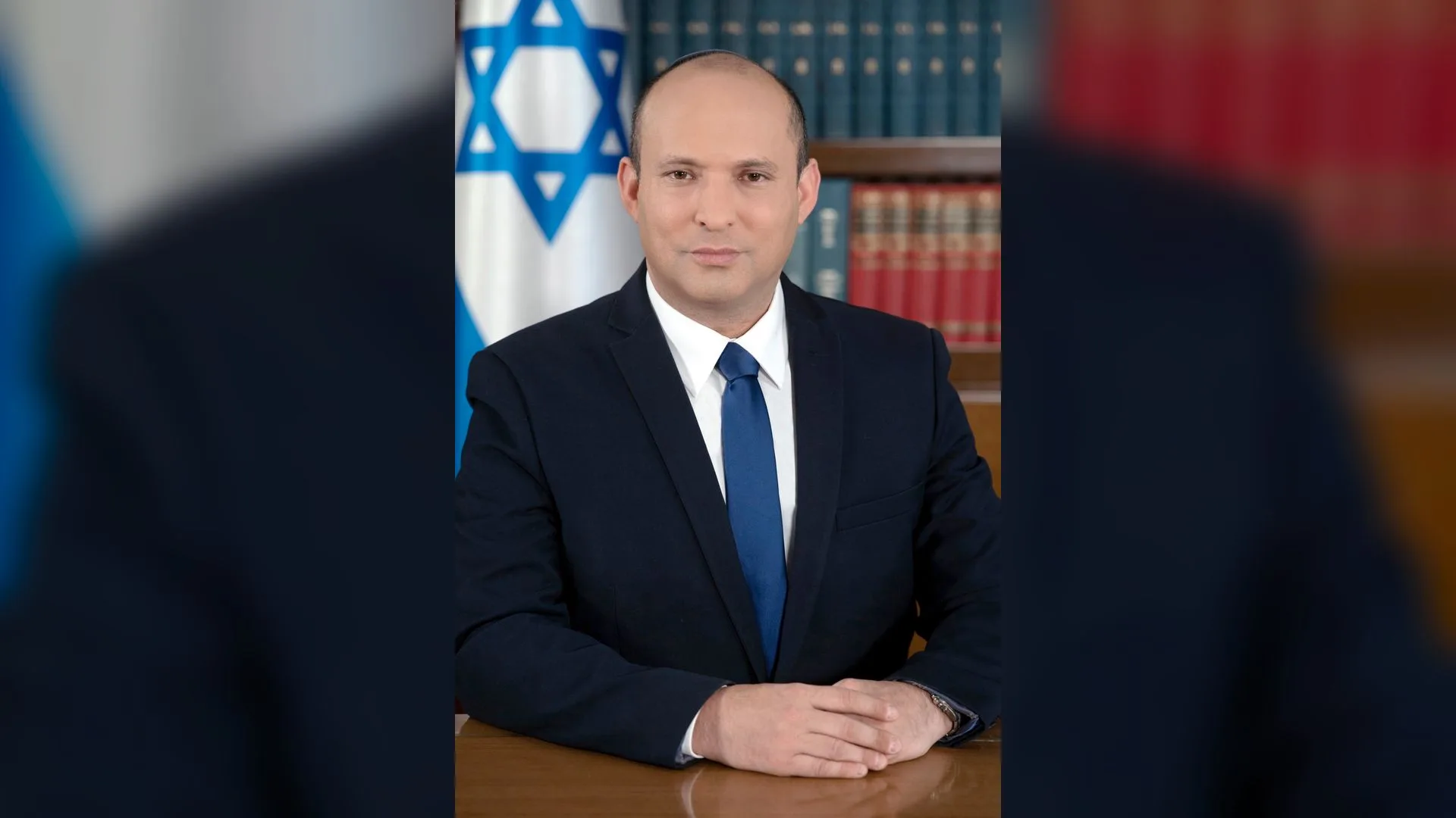 İsrail bayrağı ve kitaplık önünde masada oturan lacivert takım elbiseli, mavi kravatlı kel bir adam