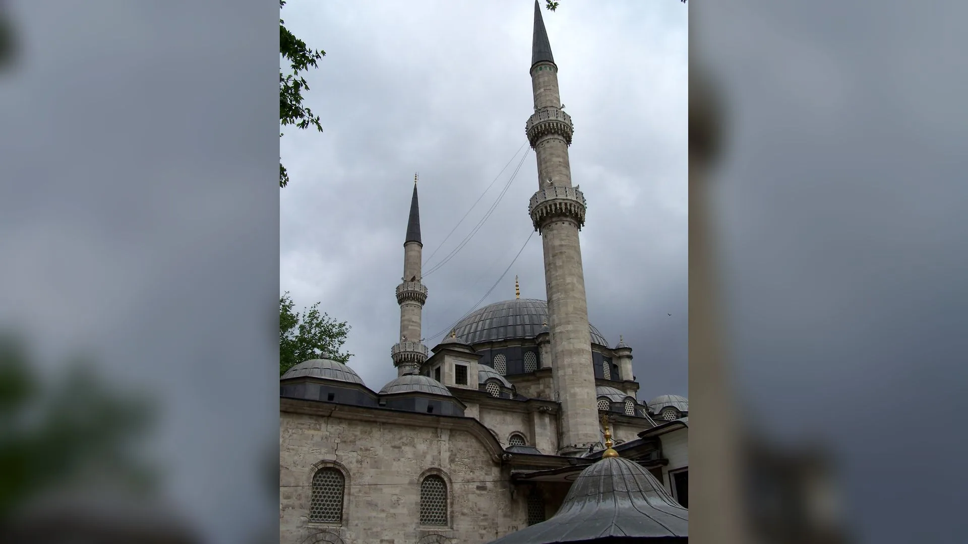 Bulutlu gökyüzü altında iki ince minareli, kurşun kubbeli taş cami