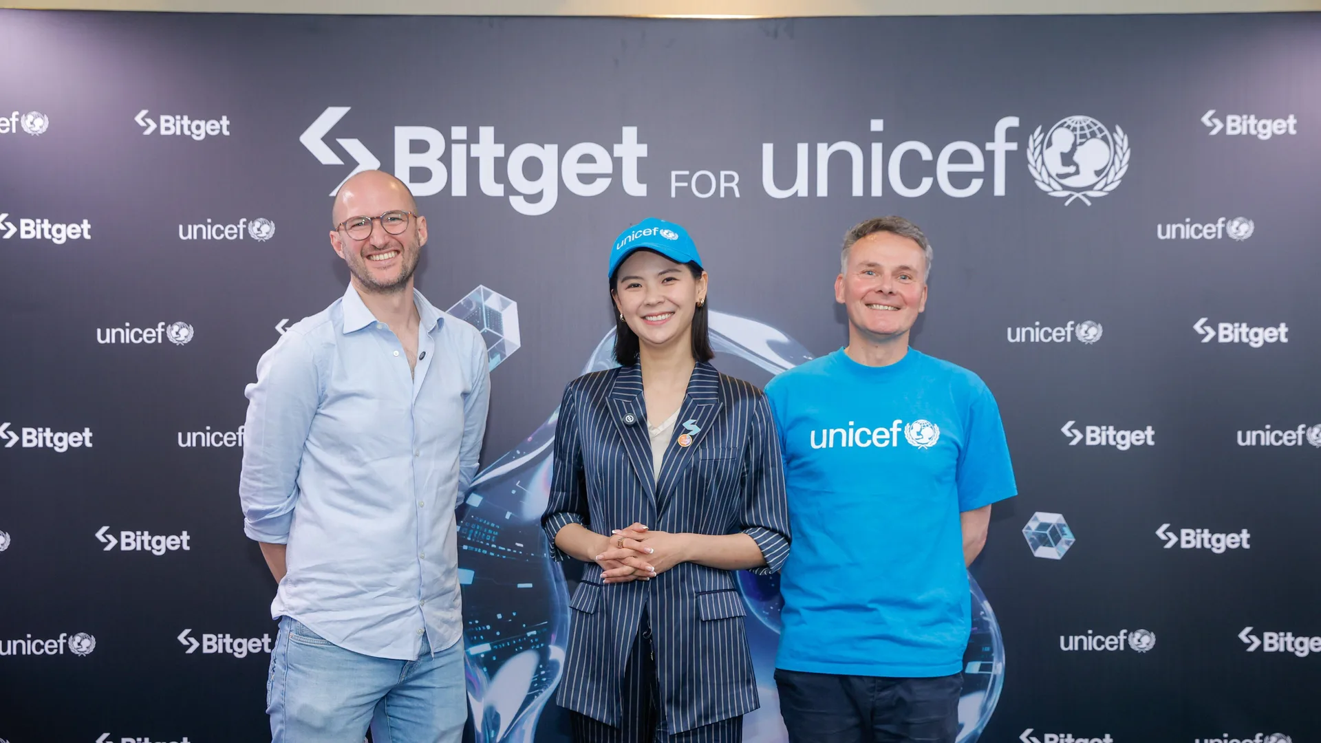 Bitget ve UNICEF logolu panonun önünde poz veren iki adam ile mavi şapkalı bir kadın