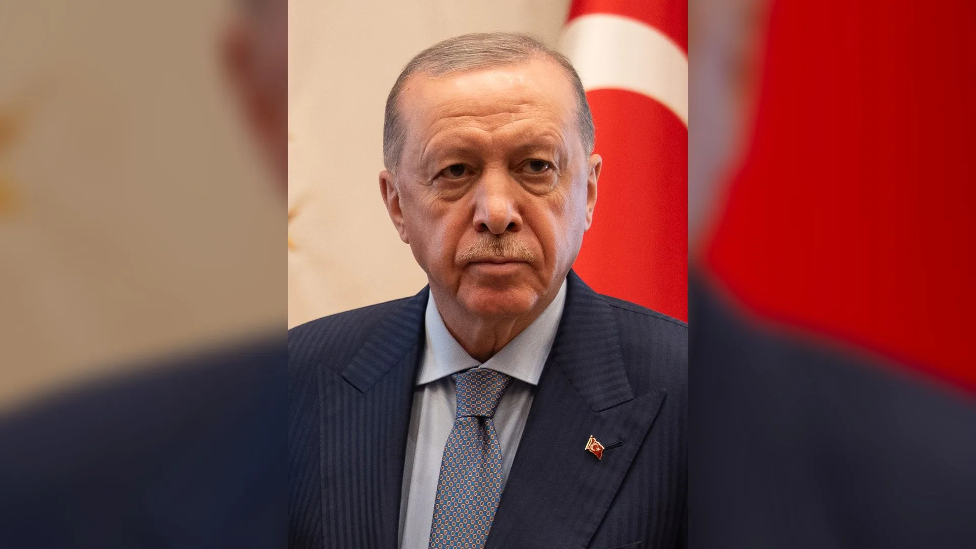 Lacivert takım elbiseli yaşlı bir adam Türk bayrağı önünde ciddi ifadeyle duruyor