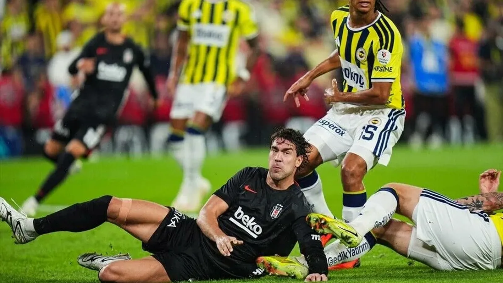 Siyah forma giymiş yerde yatan futbolcu, sarı-lacivert formadaki rakip oyuncuları etrafında.