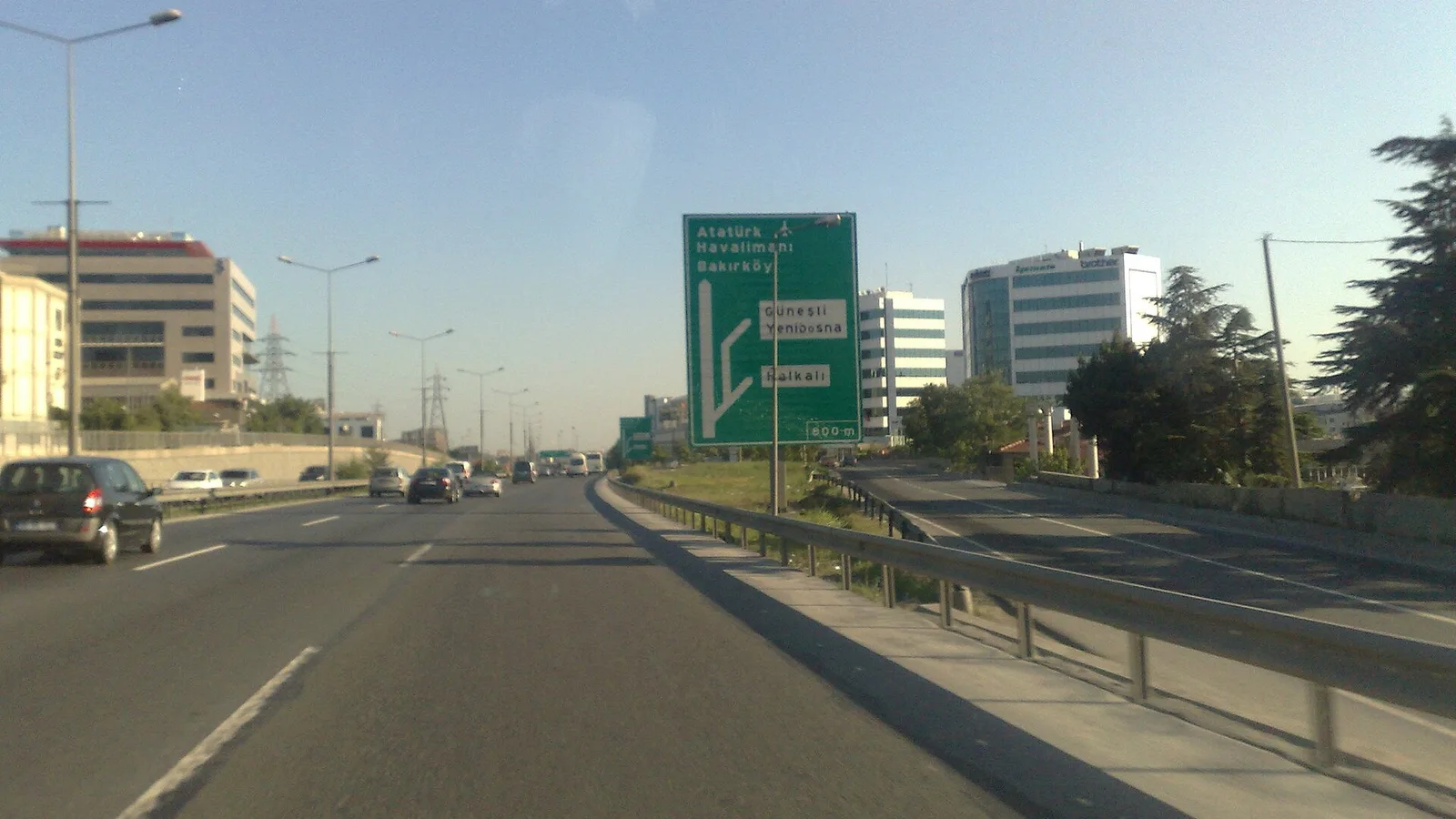 Otoyol üzerinde çok şeritli trafik akışı, yeşil işaret tabelası, şehir binaları arka planda