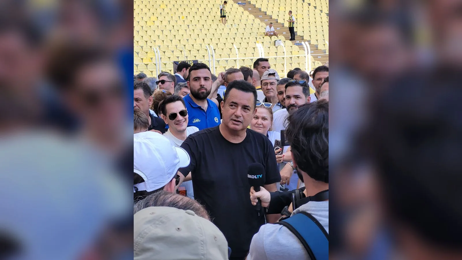 Açık havada kalabalık içinde siyah giysi giymiş adam konuşuyor.